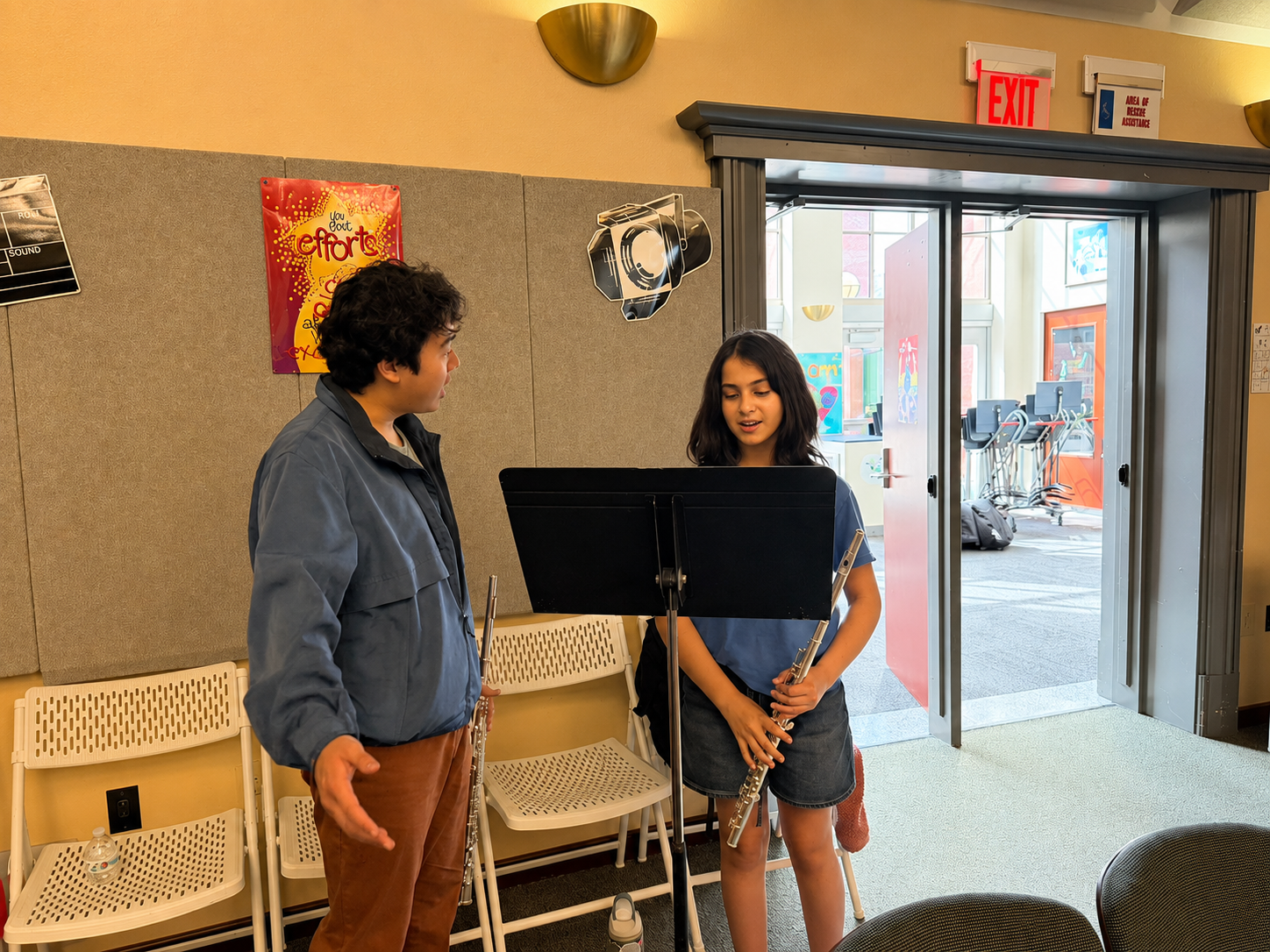Two children in a music classroom talking, one holding a flute, with a music stand and chairs behind them; door open to brightly lit art-filled room.