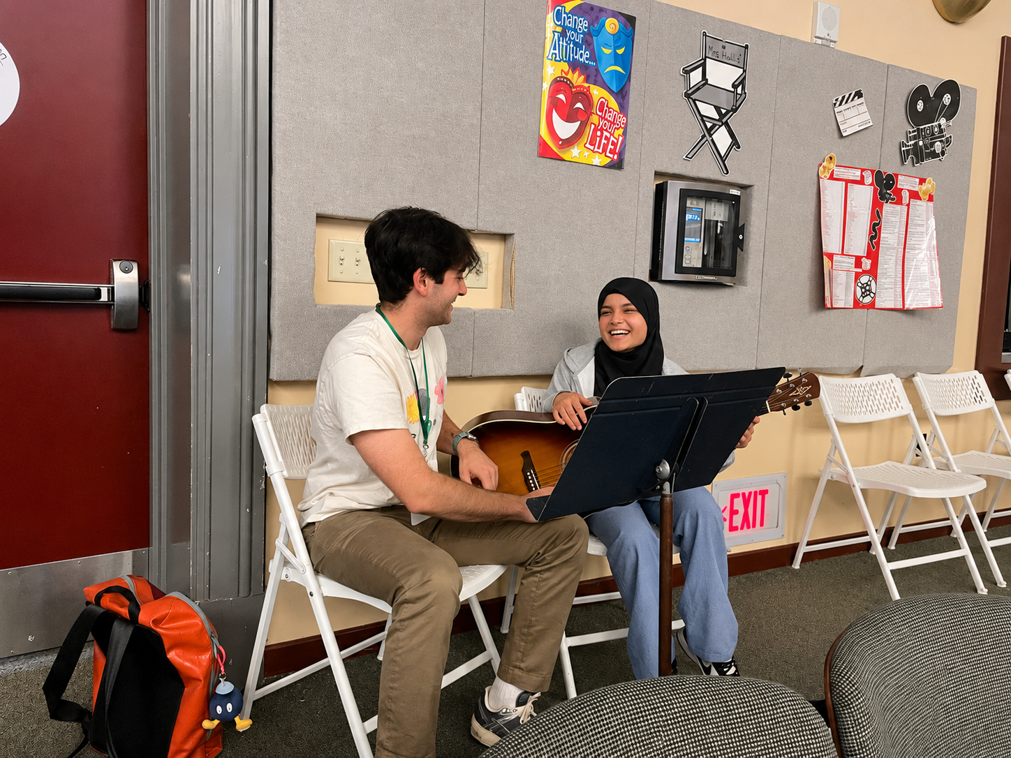 Two young people sitting on white folding chairs, smiling and playing guitar together in a hallway, with an orange backpack on the floor nearby. They are in a room with a gray wall decorated with posters and a red exit sign below them.