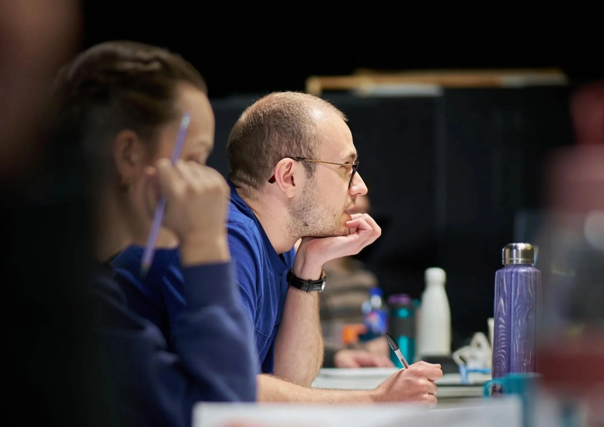 A man in glasses and a blue shirt sitting at a table with several water bottles, holding a pen and thinking.