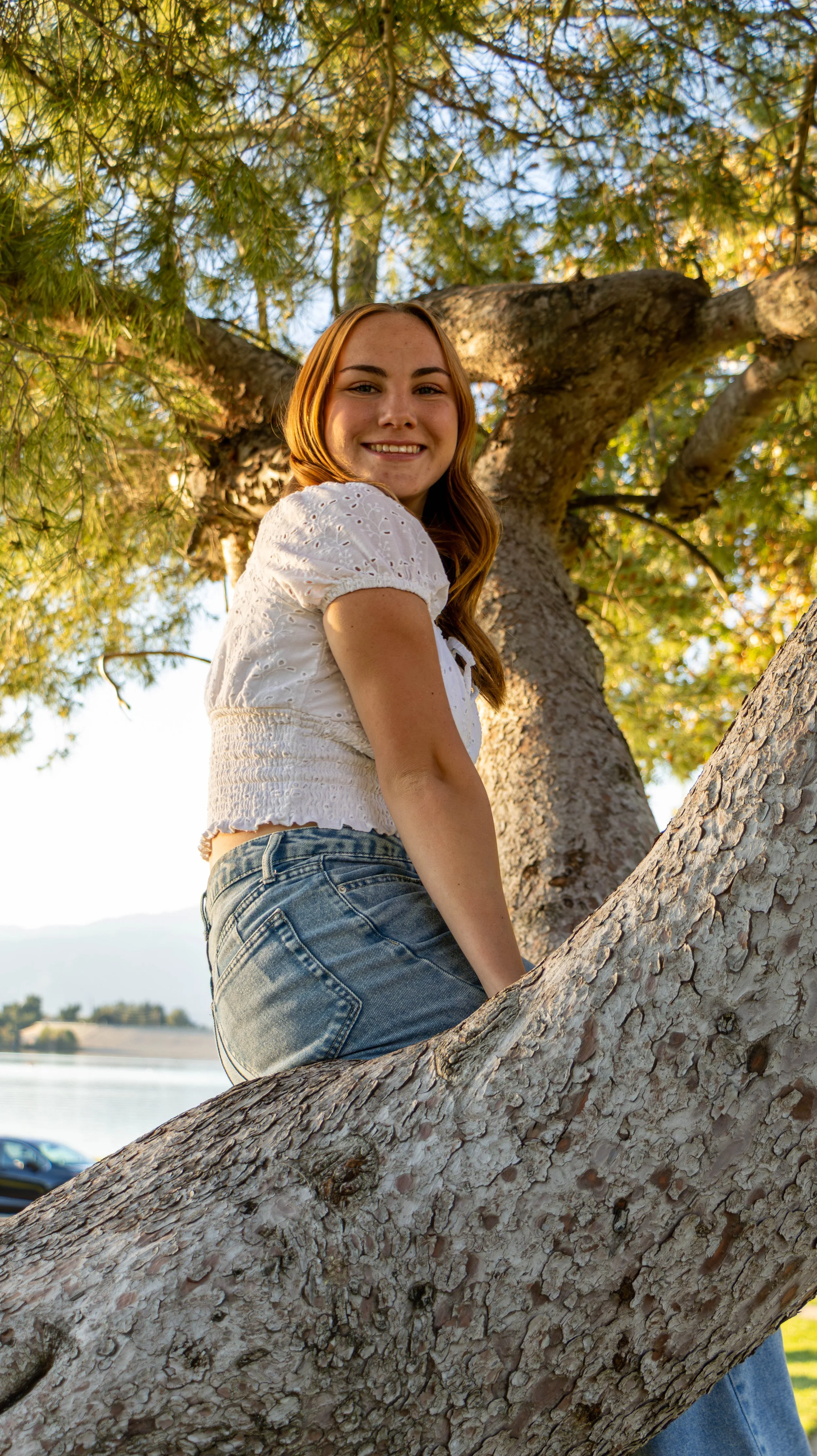 A young woman with red hair and a white eyelet blouse sitting on a large tree branch near a body of water, smiling at the camera with a scenic background of trees and sky.