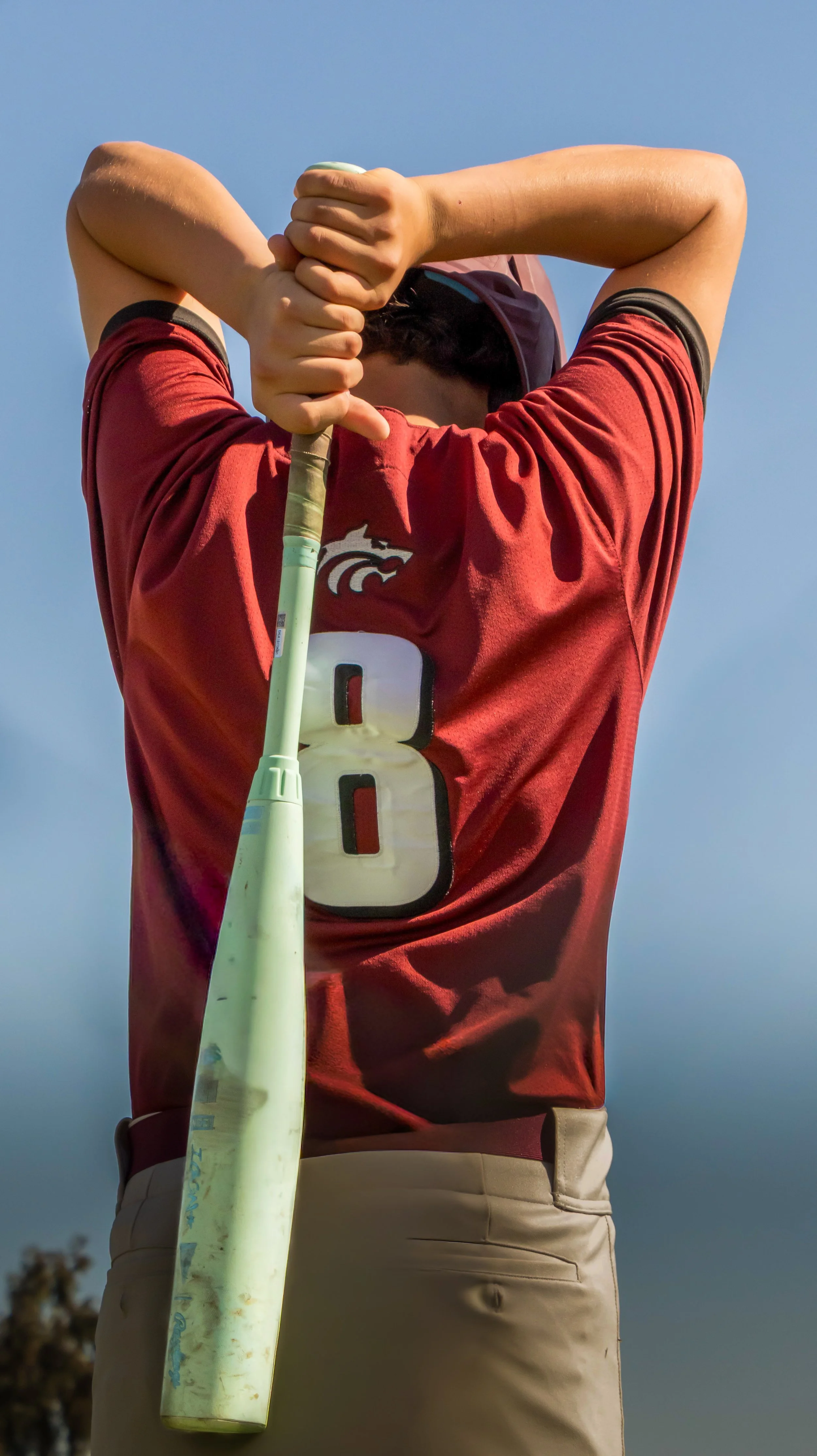 A golfer in a red shirt with a number 8 on the back, holding a golf club with both hands behind his head, standing outdoors against a clear blue sky.