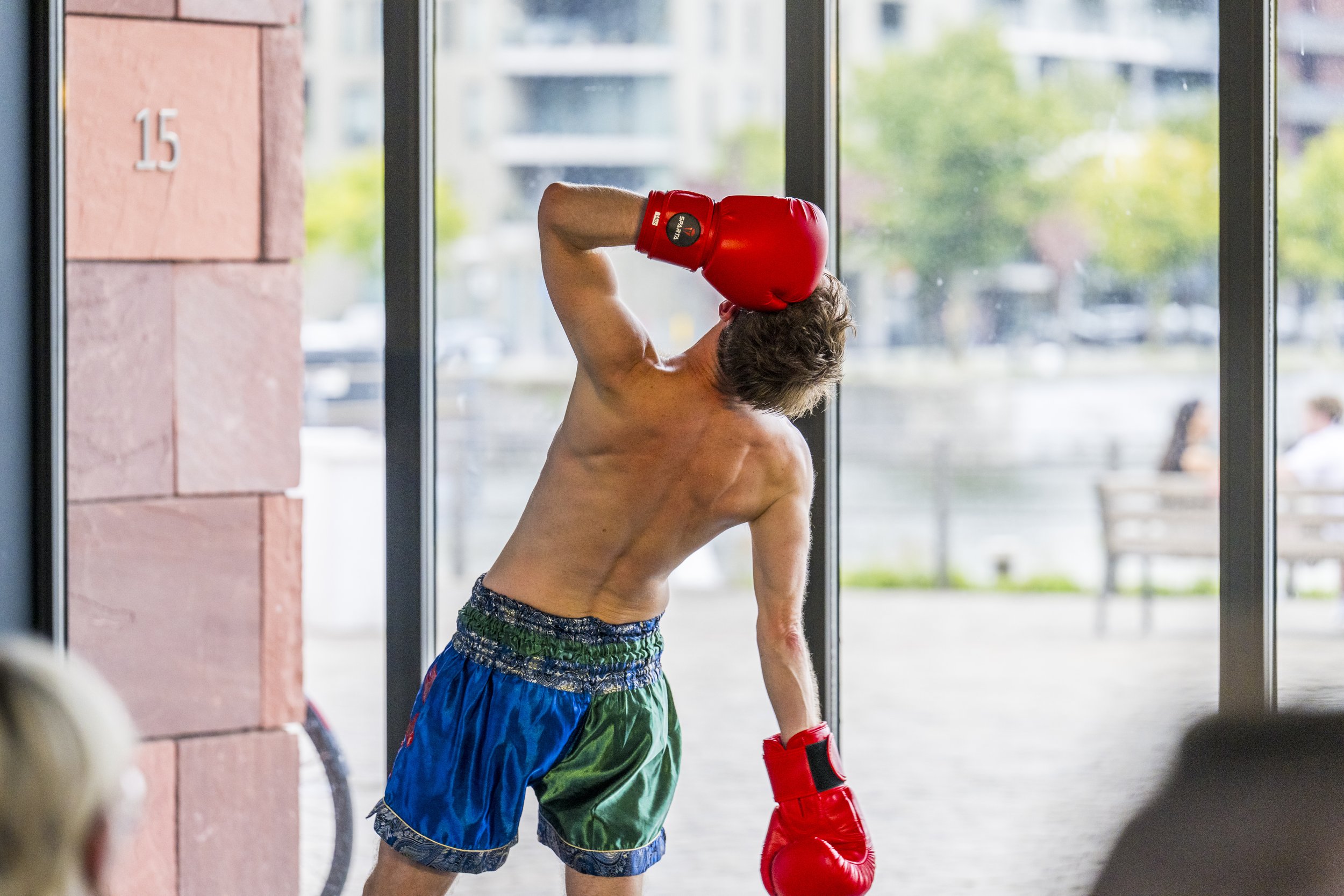 A shirtless man in colorful shorts and red boxing gloves training indoors near a large window with a city view.