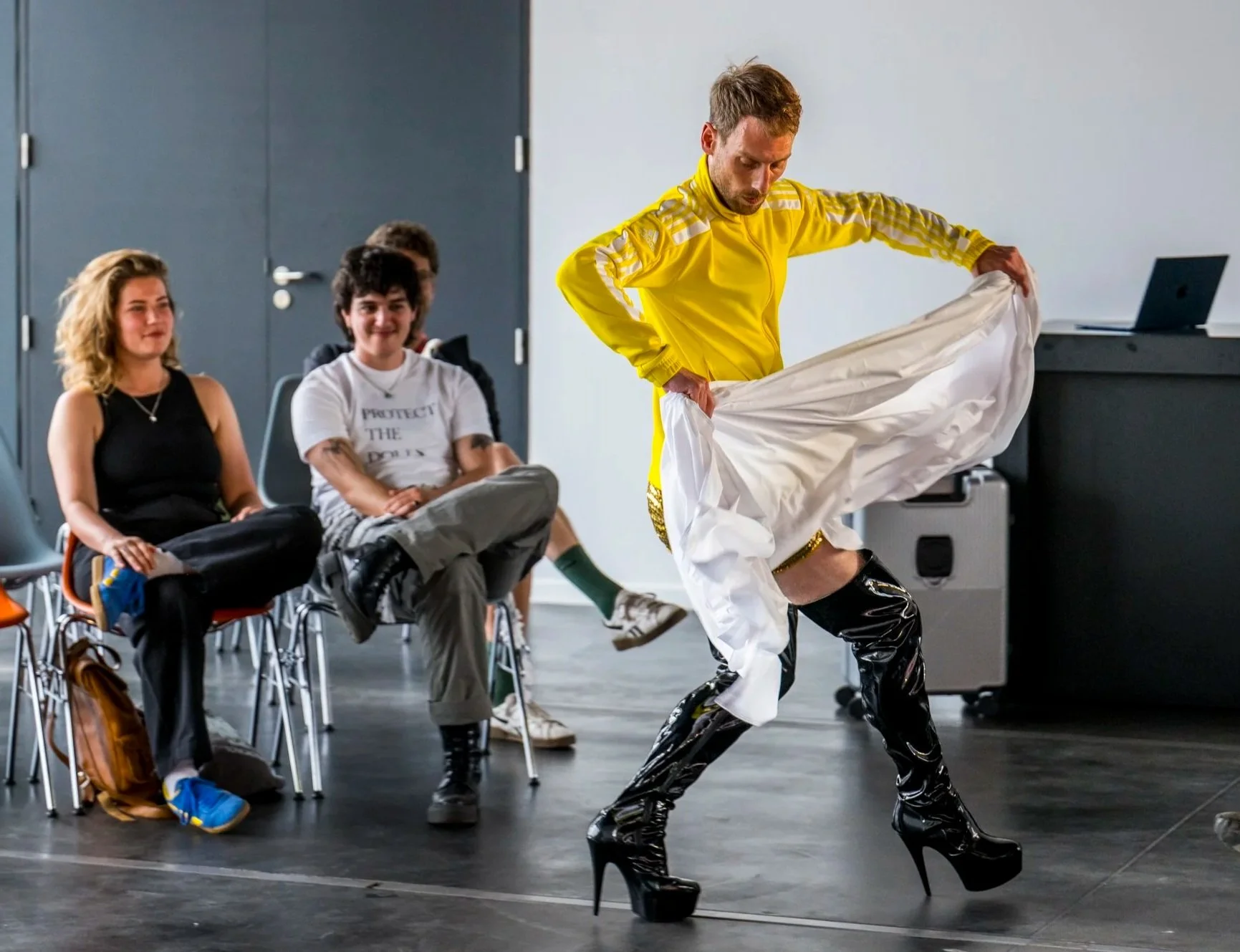 A performer in a yellow jacket with white pants and high-heeled shiny black boots in front of seated audience members watching intently.