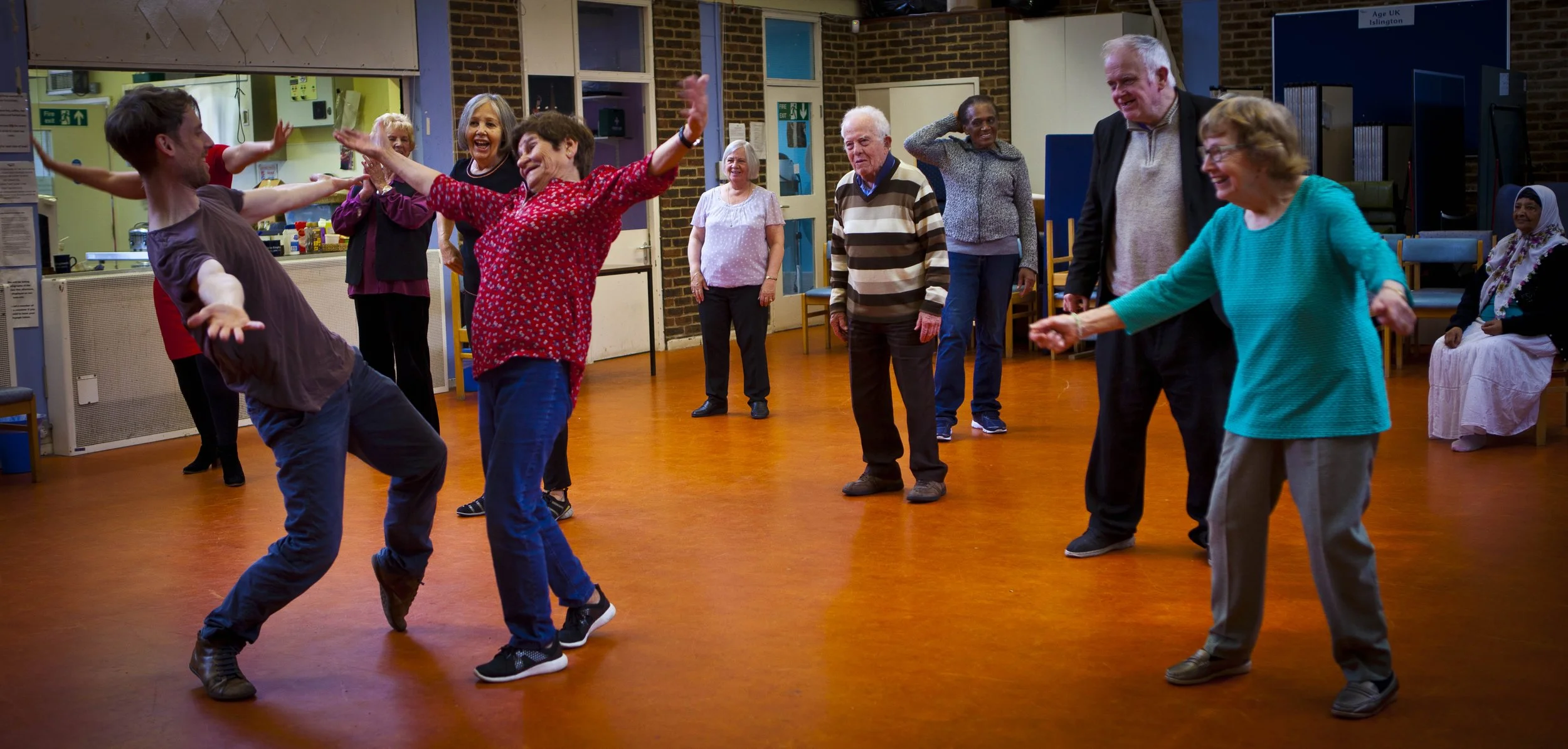 Group of elderly people dancing and laughing in a community hall with a warm wooden floor and brick walls.