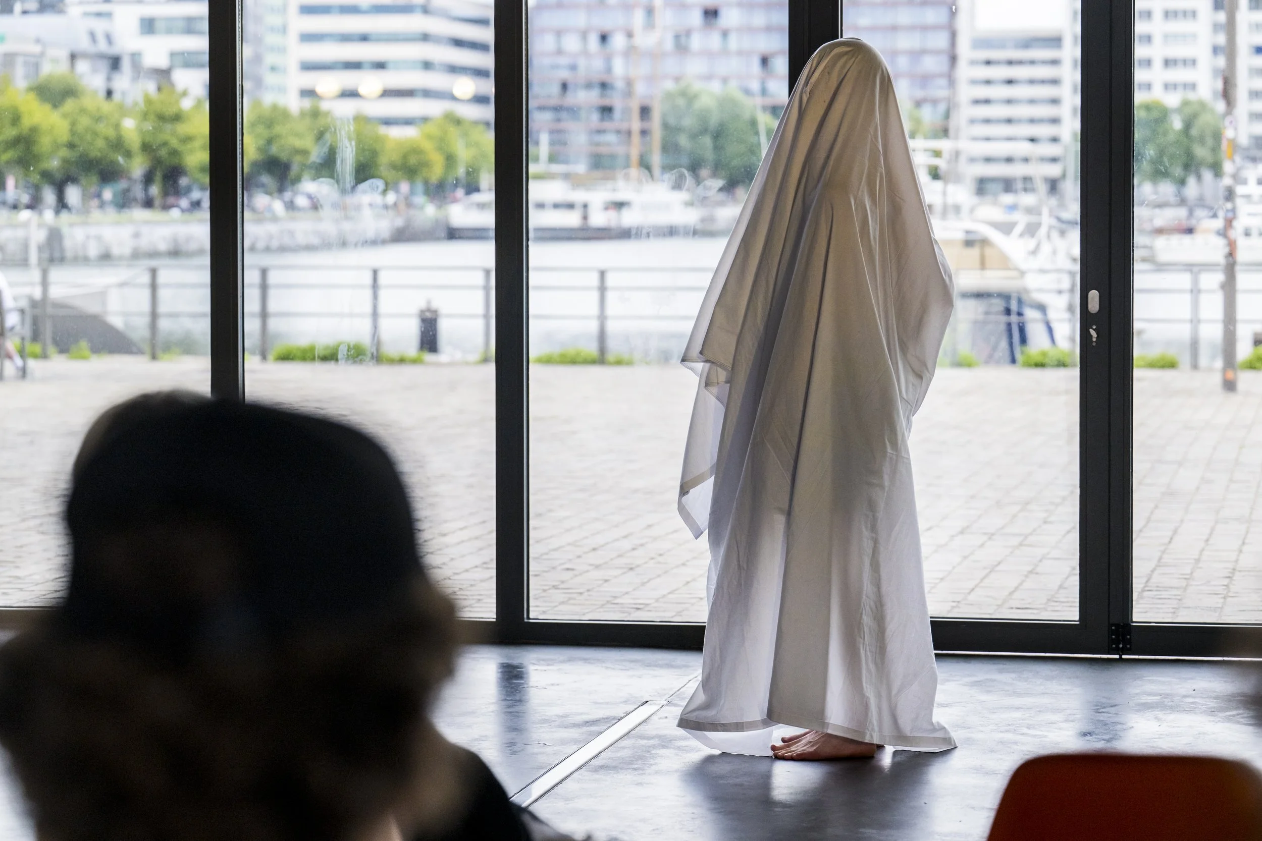 A person dressed in a long white garment called a hijab standing near a large window with a cityscape and boats in the background.