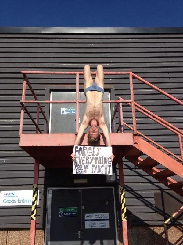 A shirtless man with shorts is performing a handstand upside down on a metal platform, holding a sign that says "FORGET EVERYTHING YOU'VE BEEN TAUGHT" in front of a building with a grey exterior and blue sky.