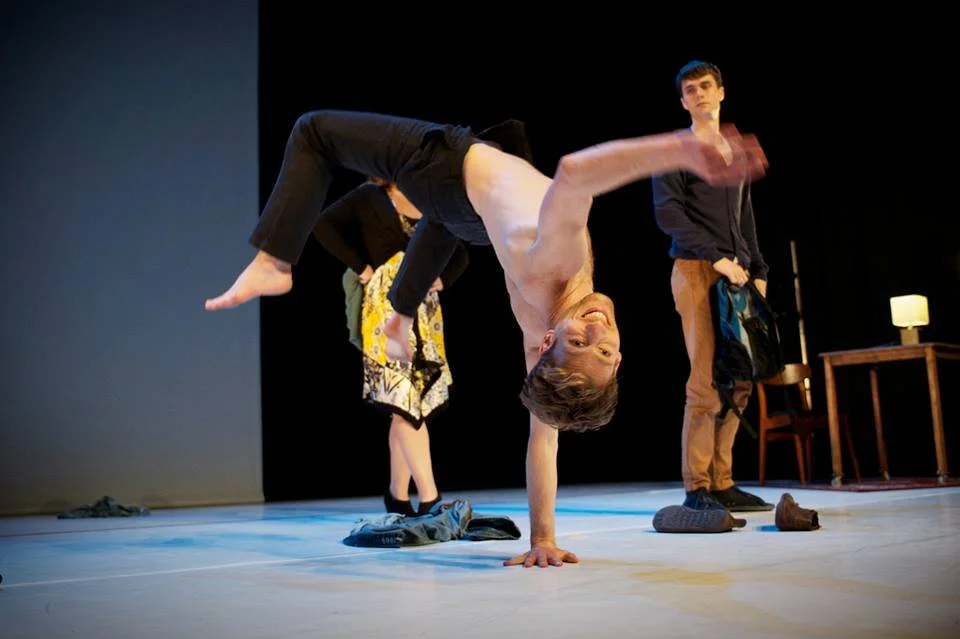 A shirtless man performing a handstand on a stage, with others in the background and stage props visible.