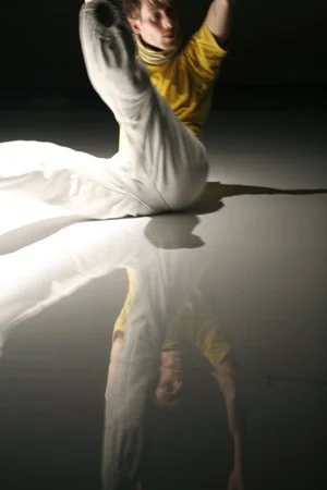 A young person in a yellow shirt and white pants sitting on the floor, with their arm raised and looking forward, casting a shadow on the surface.