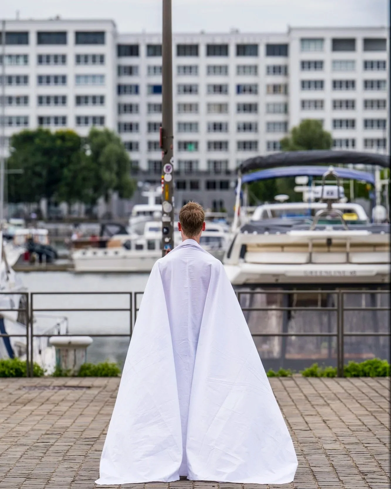 A man with short hair standing on a waterfront promenade, wearing a long white garment, looking out onto a marina with boats and a large white building in the background.