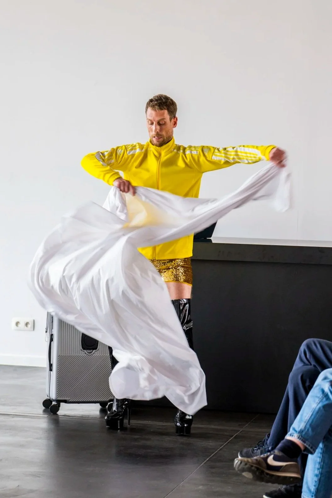 Man dressed in a bright yellow jacket, gold sequined shorts, and high platform boots performs a dance with a large white sheet in an indoor setting, with a black counter and audience members in jeans visible.