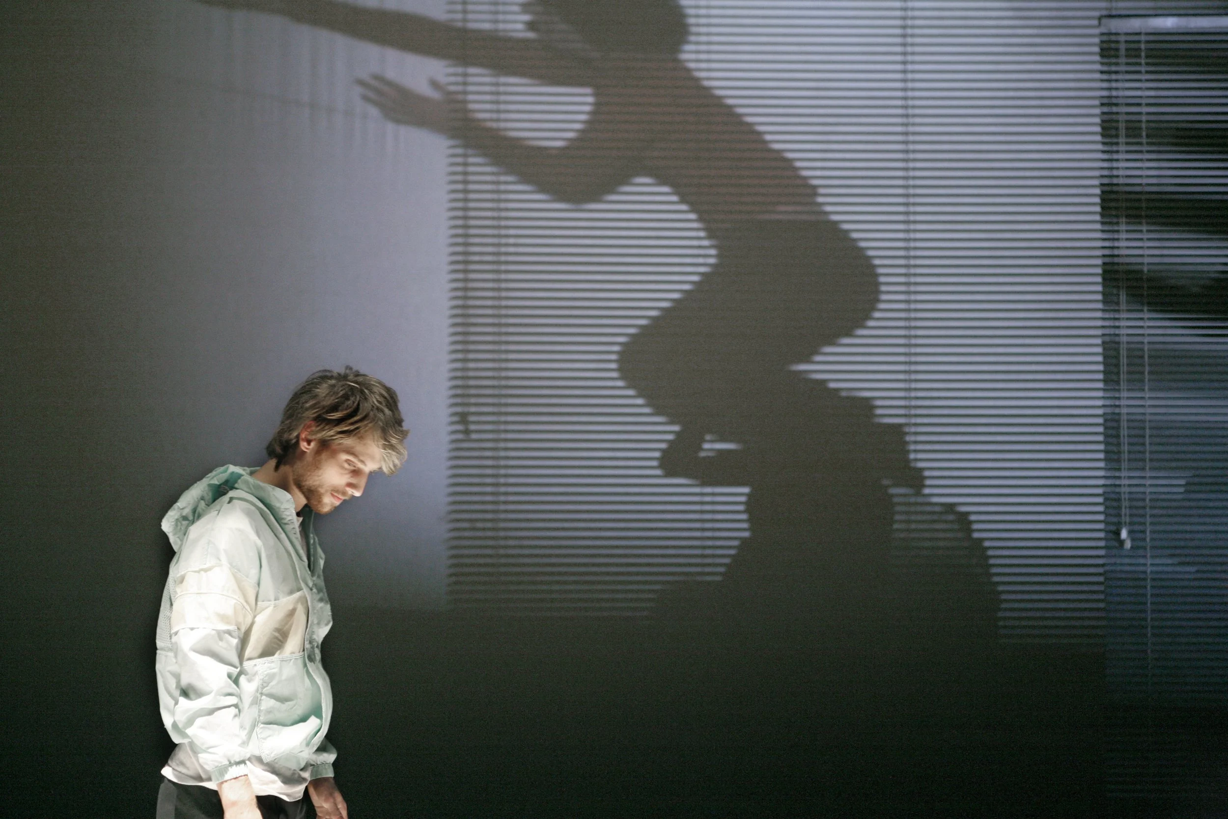 A man with brown hair and a beard looking down, wearing a light-colored jacket, standing against a dark wall with horizontal blinds and a large shadow of a seated person cast on the wall.