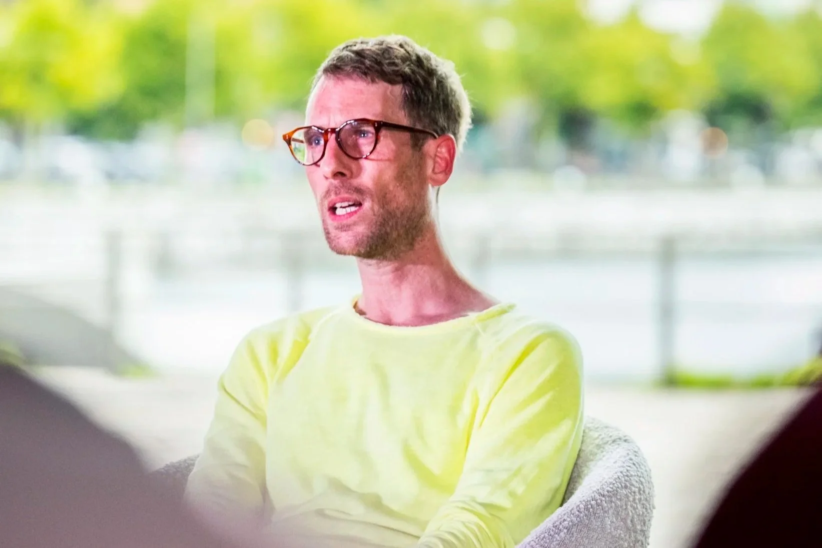 A man wearing glasses and a yellow shirt is sitting outdoors with a blurred background of trees and a railing.