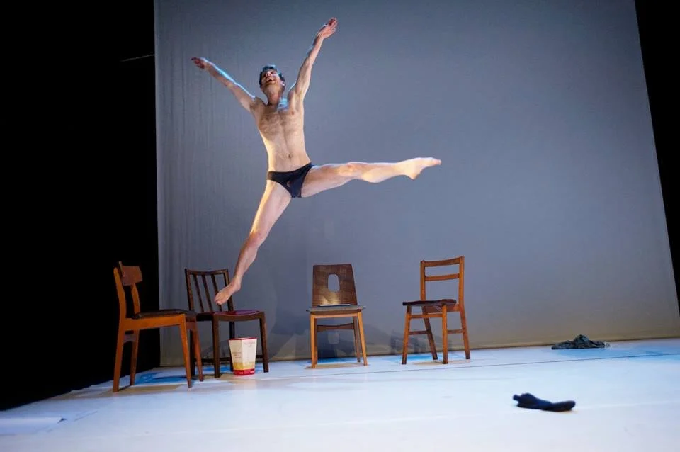A man mid-air during a jump on a stage with three chairs, a bucket, and some clothes on the floor.