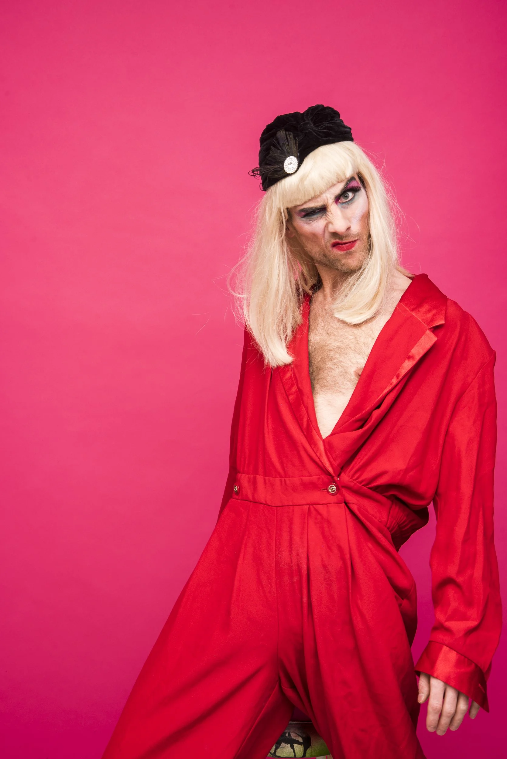 A person dressed in pink from head to toe, wearing a black turban with a decorative brooch, has long blonde hair, heavy makeup, and is standing against a bright pink wall.