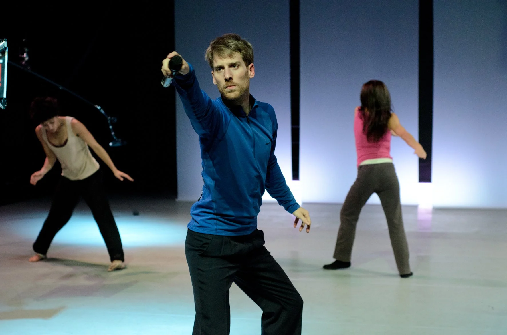 A man in a blue long-sleeve shirt and black pants holding a microphone appears to be leading a dance rehearsal or class, with two women in the background also dancing; one wearing a white tank top and black pants, the other wearing a pink tank top an