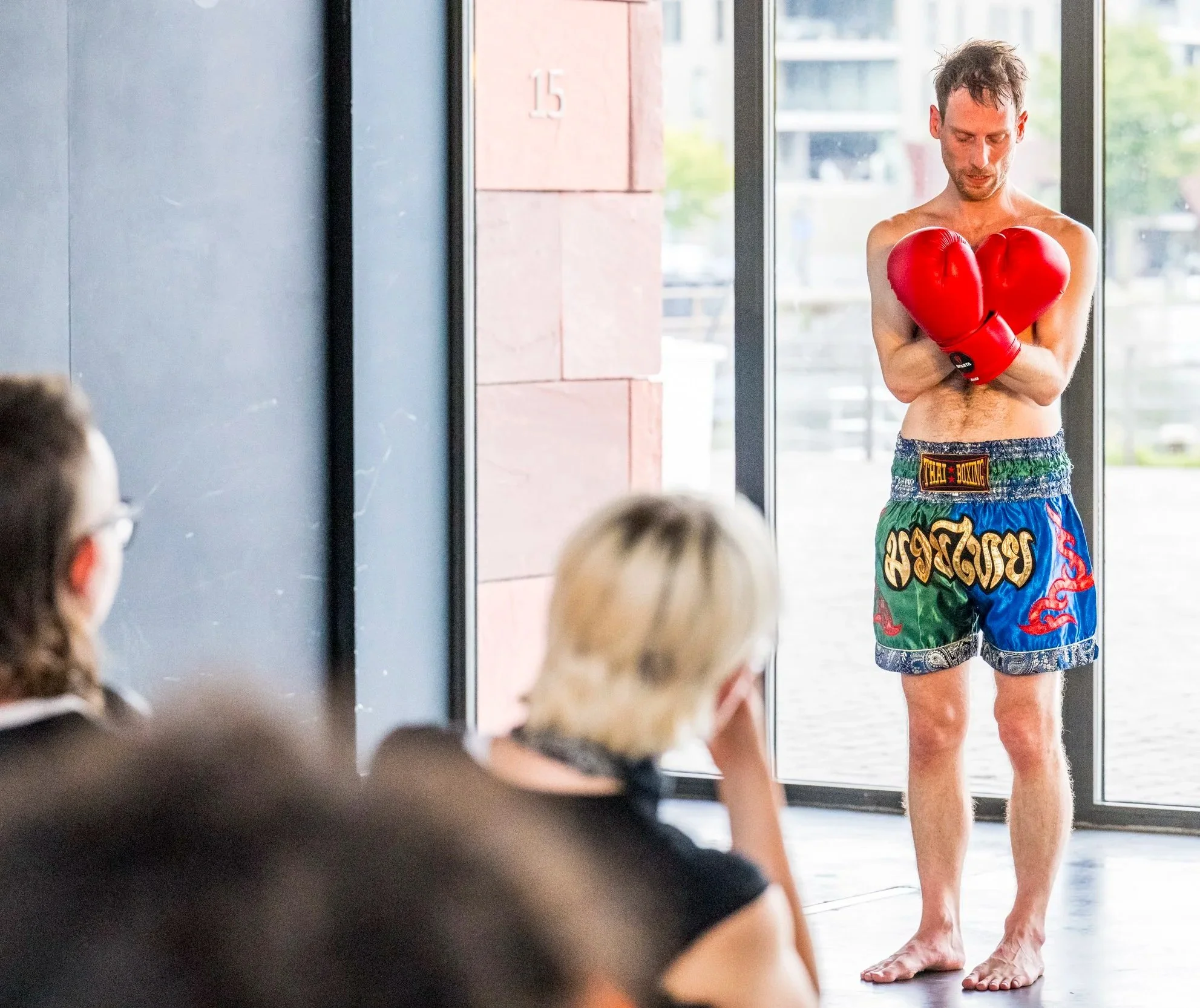 A shirtless man wearing boxing shorts and red boxing gloves standing in front of a glass door, looking down with a serious expression, with a blurred group of children sitting inside watching him.