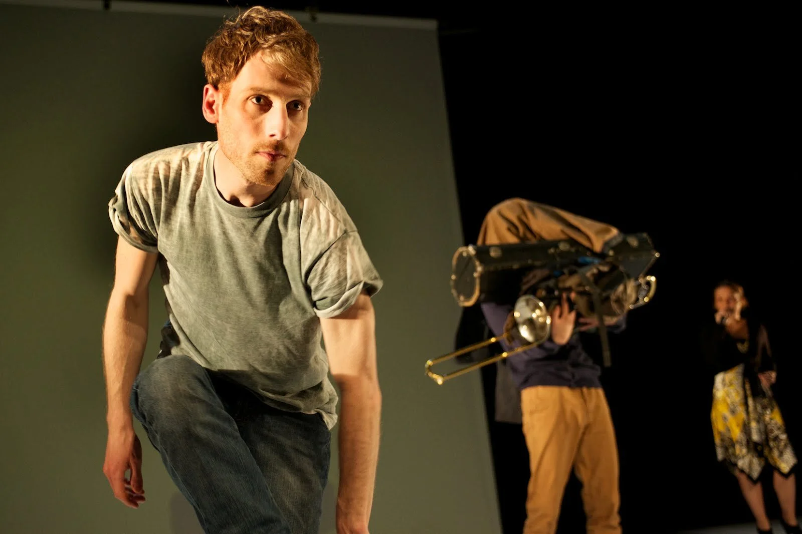 A man with curly red hair, wearing a gray tie-dye t-shirt and dark jeans, is leaning forward on a stage with a serious expression. Behind him, a person with a trombone on their back and another woman are present, suggesting a theater or performance s