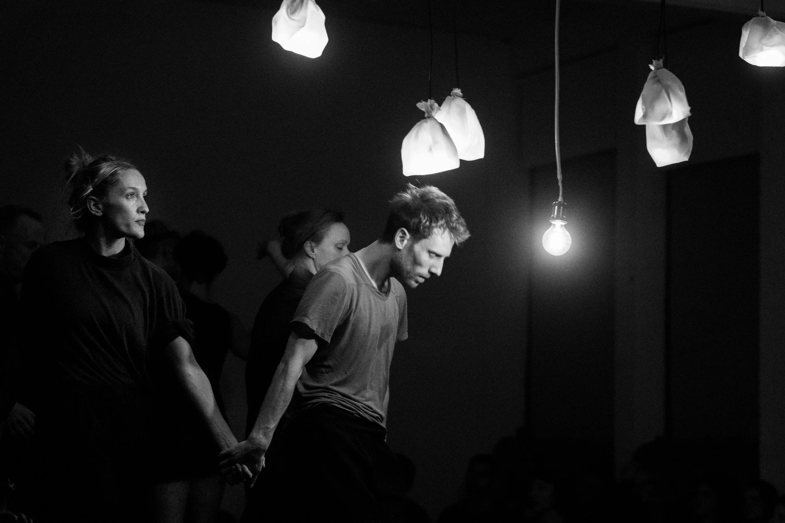 Black and white photo of four people standing in a dimly lit room with hanging light bulbs and fabric-covered light fixtures. They seem to be deep in thought or concentration.