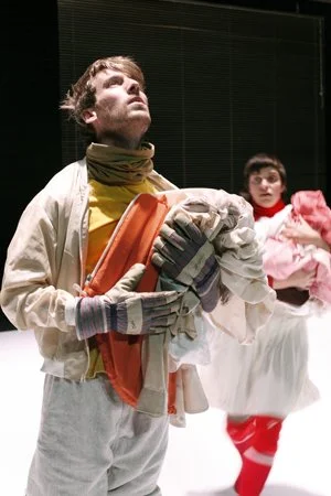 A young man dressed in a yellow shirt, beige jacket, and gray gloves, holding clothes and a box, looking upward, with a woman in a pink dress in the background.