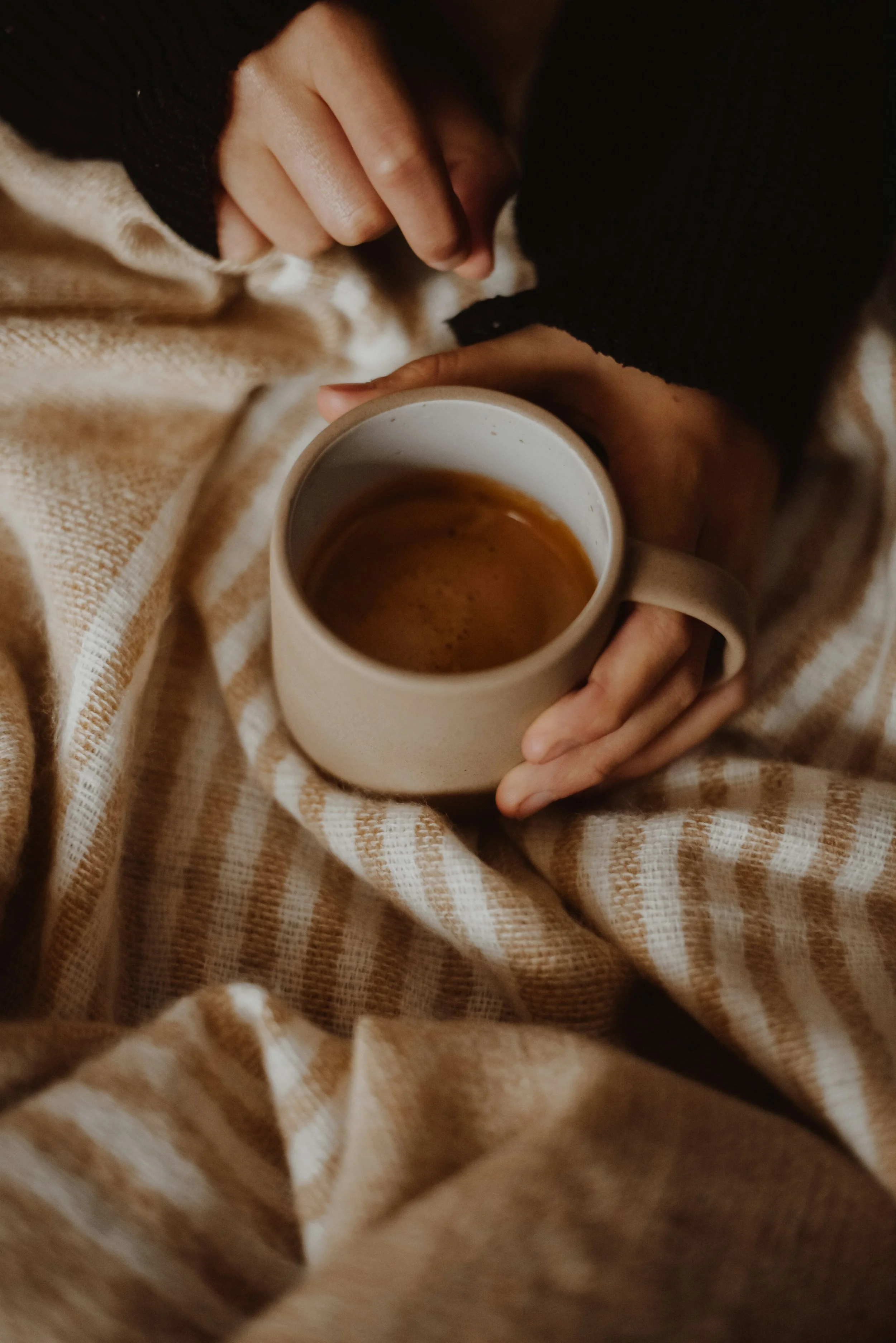 A person holding a ceramic mug with a hot beverage, sitting on a beige and white striped blanket.