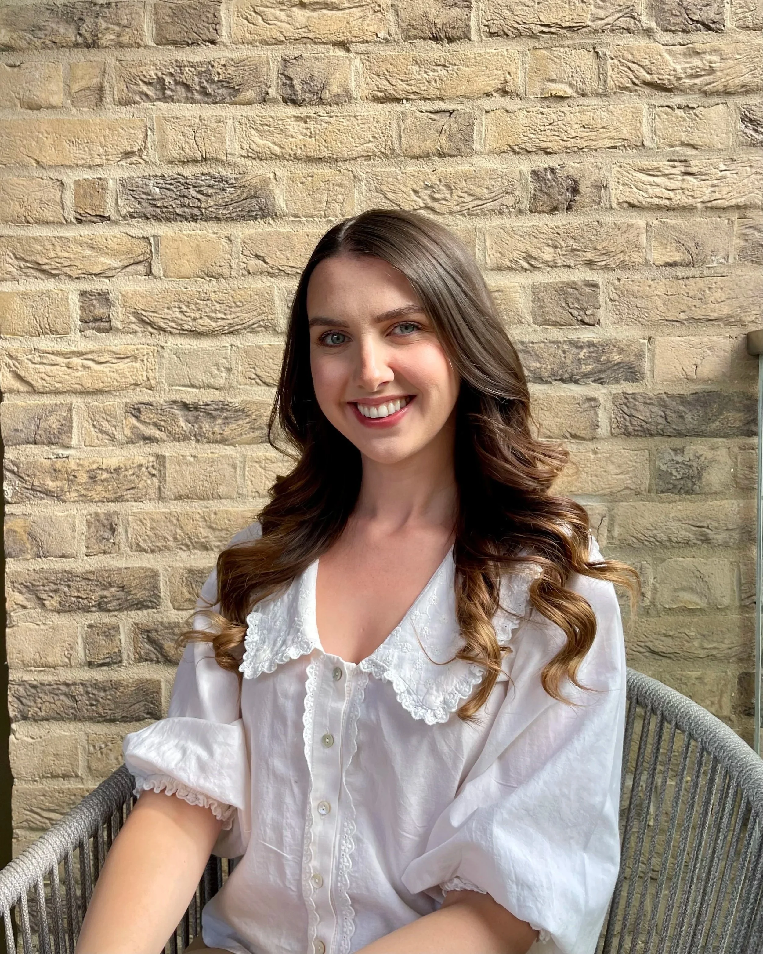 A female therapist and psychologist with long brown hair and blue eyes sitting on a metal chair with gray woven pattern. She is smiling and wearing a white blouse with lace details around the collar and sleeves. She is in front of a beige-brick wall.