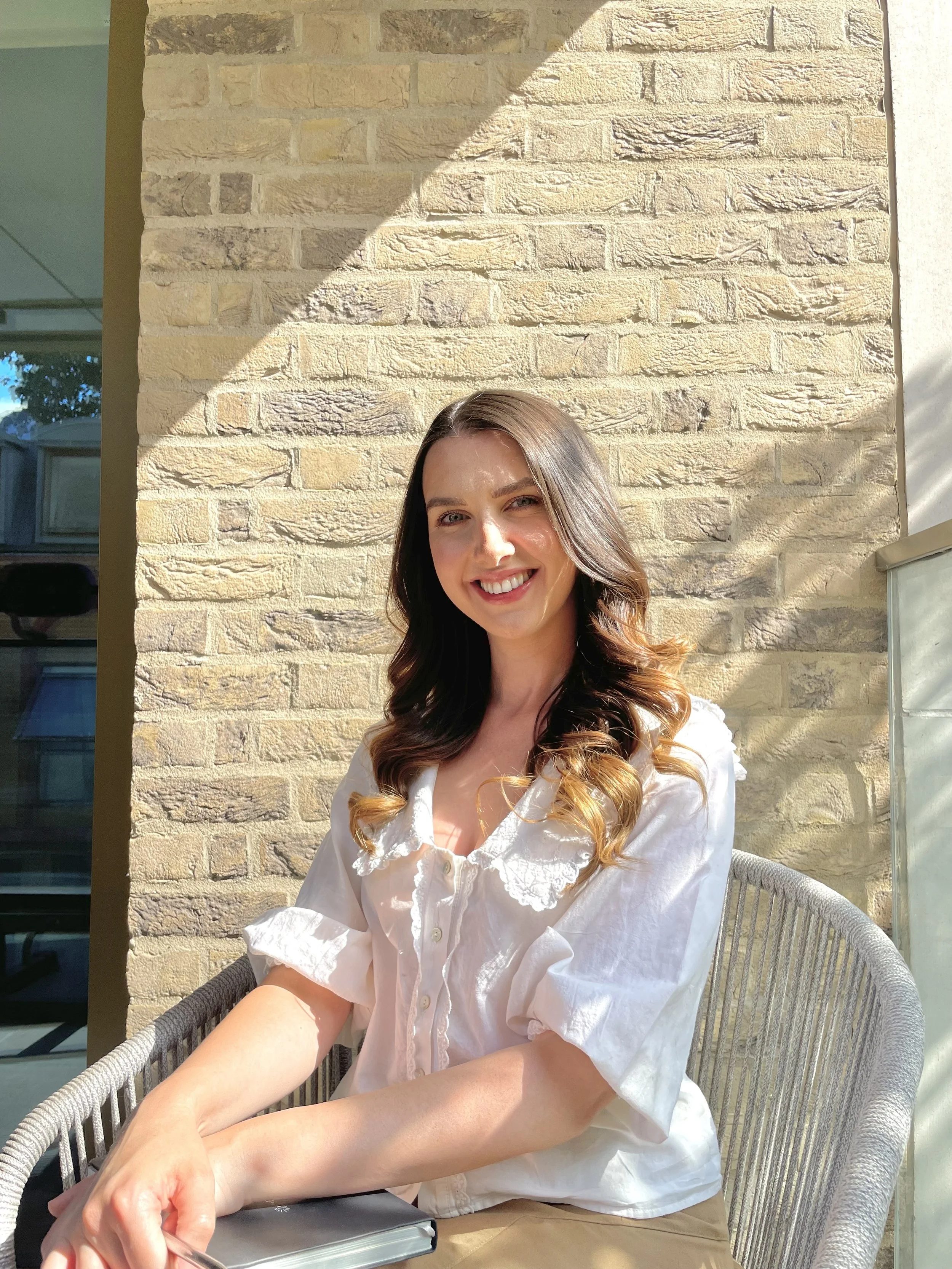 A woman with daylights and curled hair sitting outdoors on a patio, smiling, with a beige brick wall behind her and sunlight casting shadows.