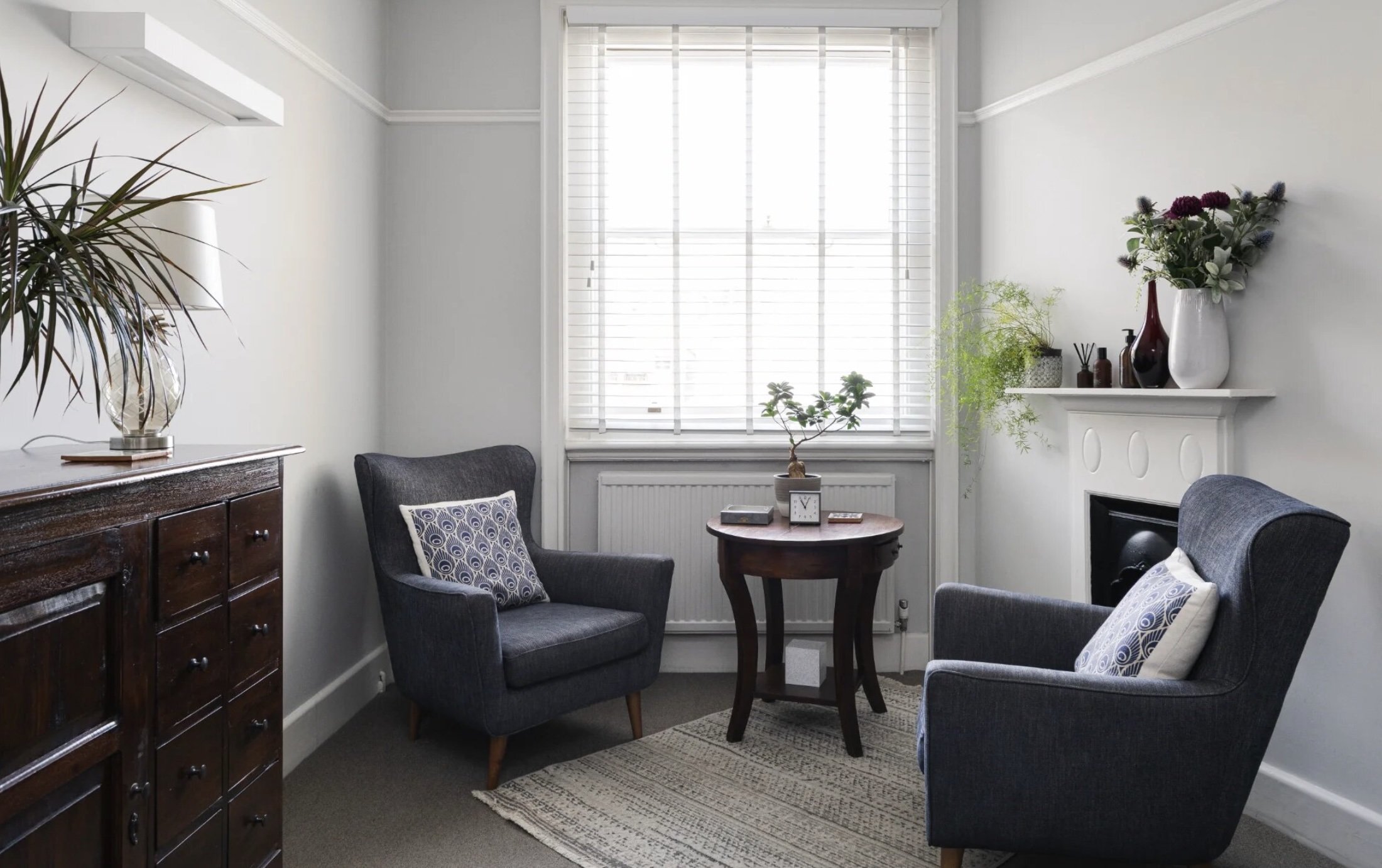 A cozy living room with a window covered by white blinds, a small round wooden table with a potted plant, a clock, and a book, flanked by two gray armchairs with patterned pillows. There's a dark wooden sideboard on the left with a large plant in a glass vase. On the right, a white mantel with vases and decorative items next to a fireplace. A small rug is on the floor, and the walls are painted light gray.