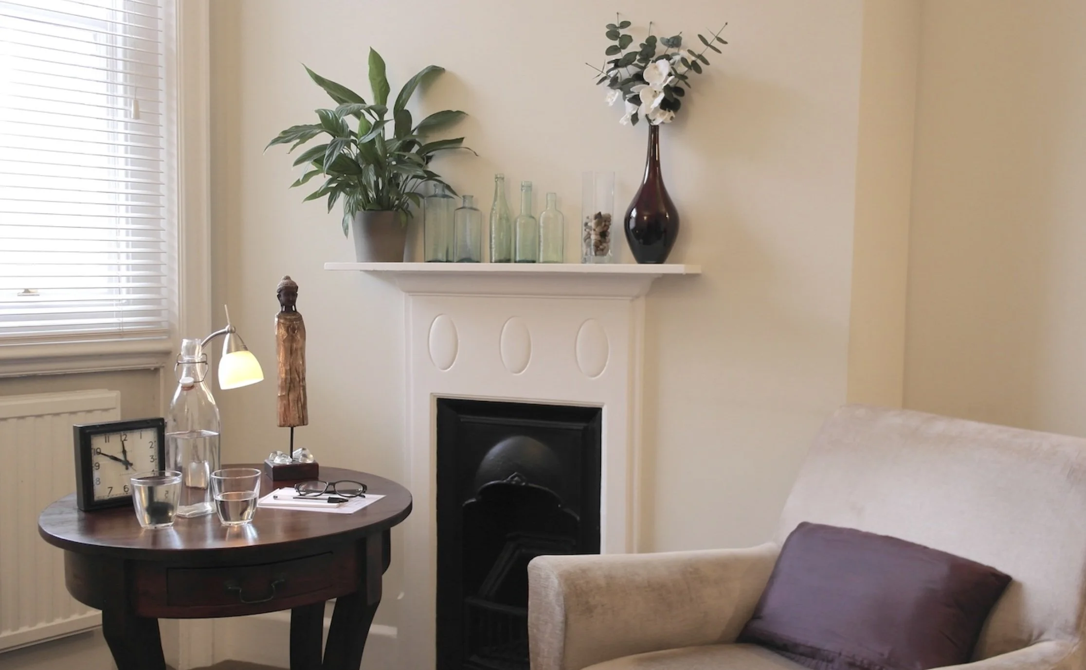 A living room corner with a beige armchair, a round wooden table with glasses, a clock, and papers, a lamp, a fireplace with a mantel decorated with potted plants and vases, a window with blinds, and a portion of a couch.