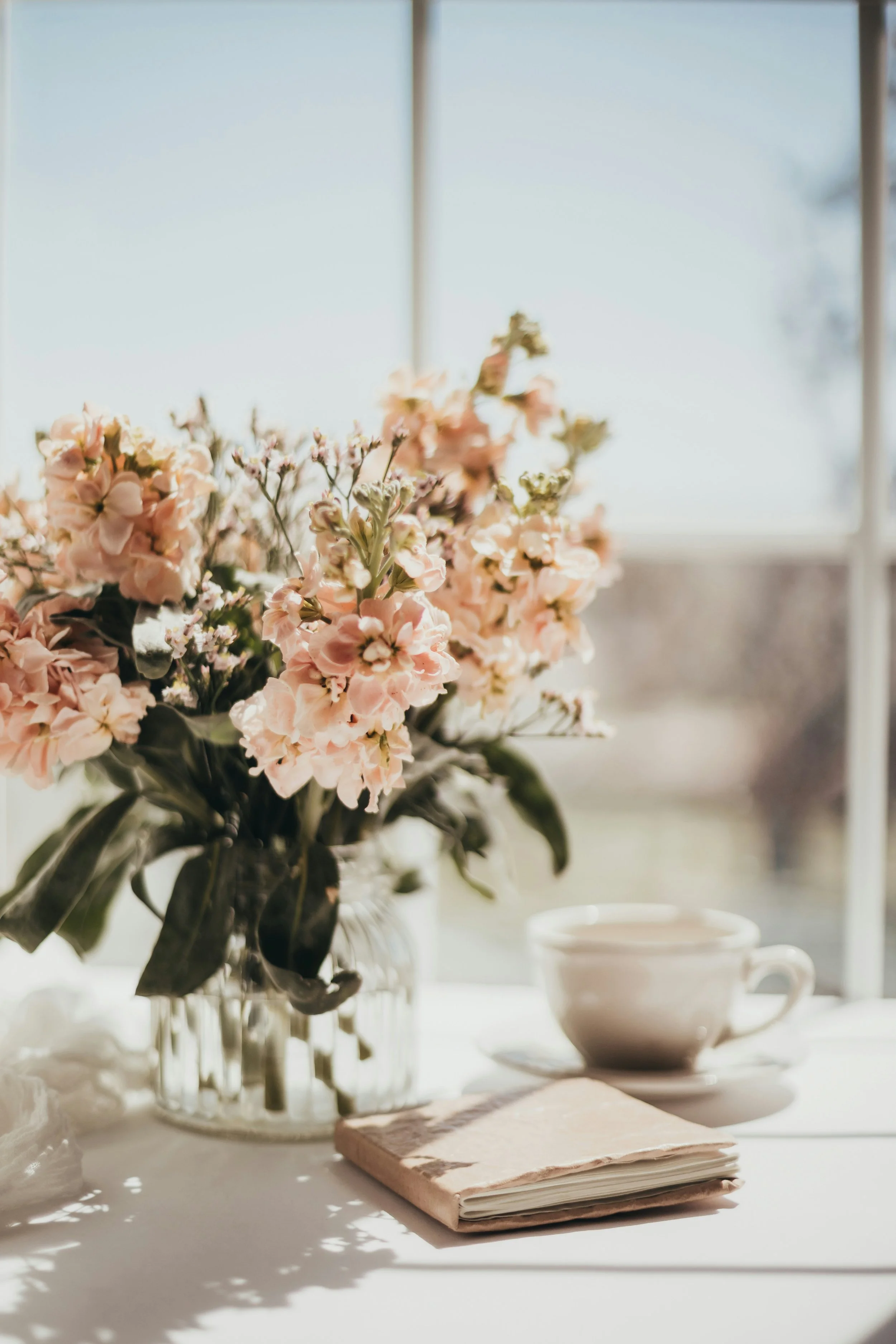 A bouquet of pink and white flowers in a glass vase on a white table with a white coffee cup and a closed notebook nearby, in front of a window with sunlight streaming in.