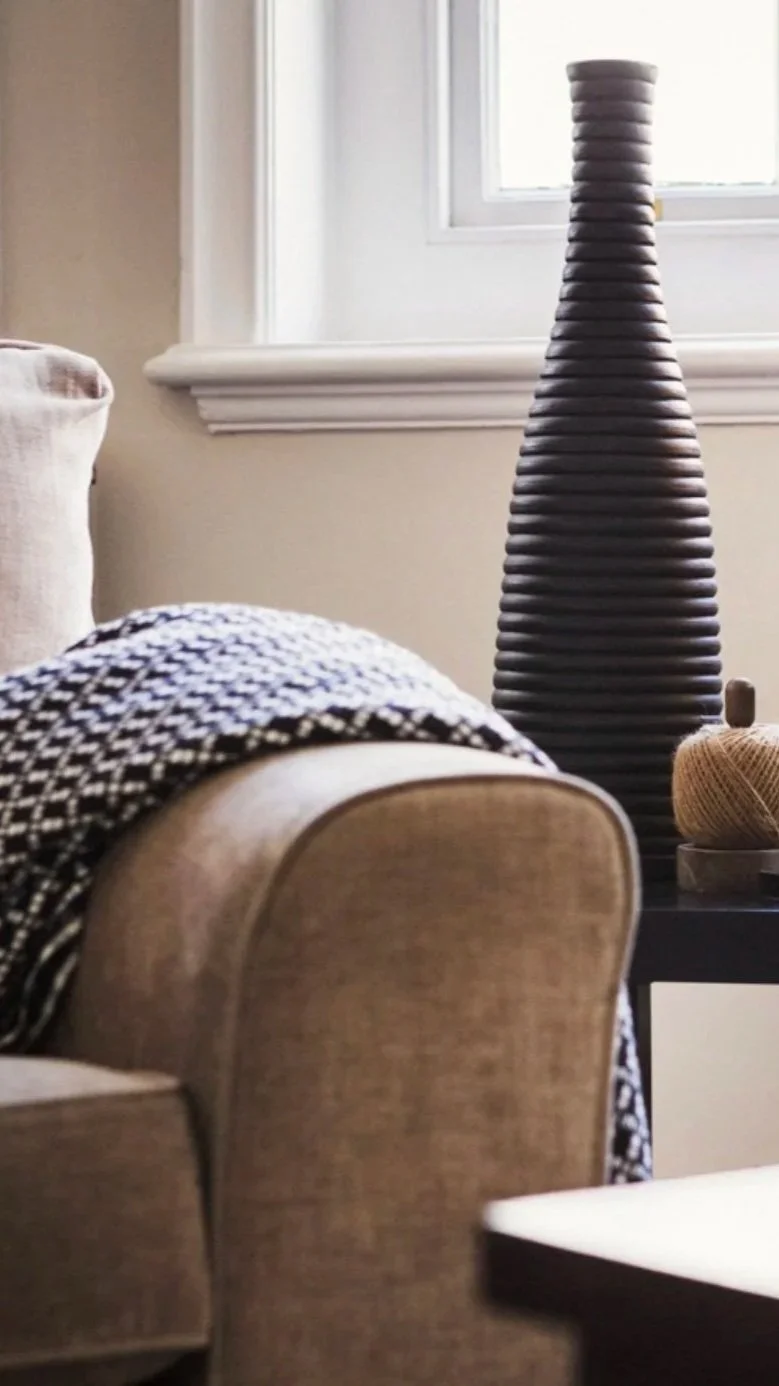 A living room corner featuring a beige upholstered chair, a black and white patterned throw blanket, a tall dark ribbed ceramic vase, a window with white trim, and part of a small side table with a yellow knitted object.
