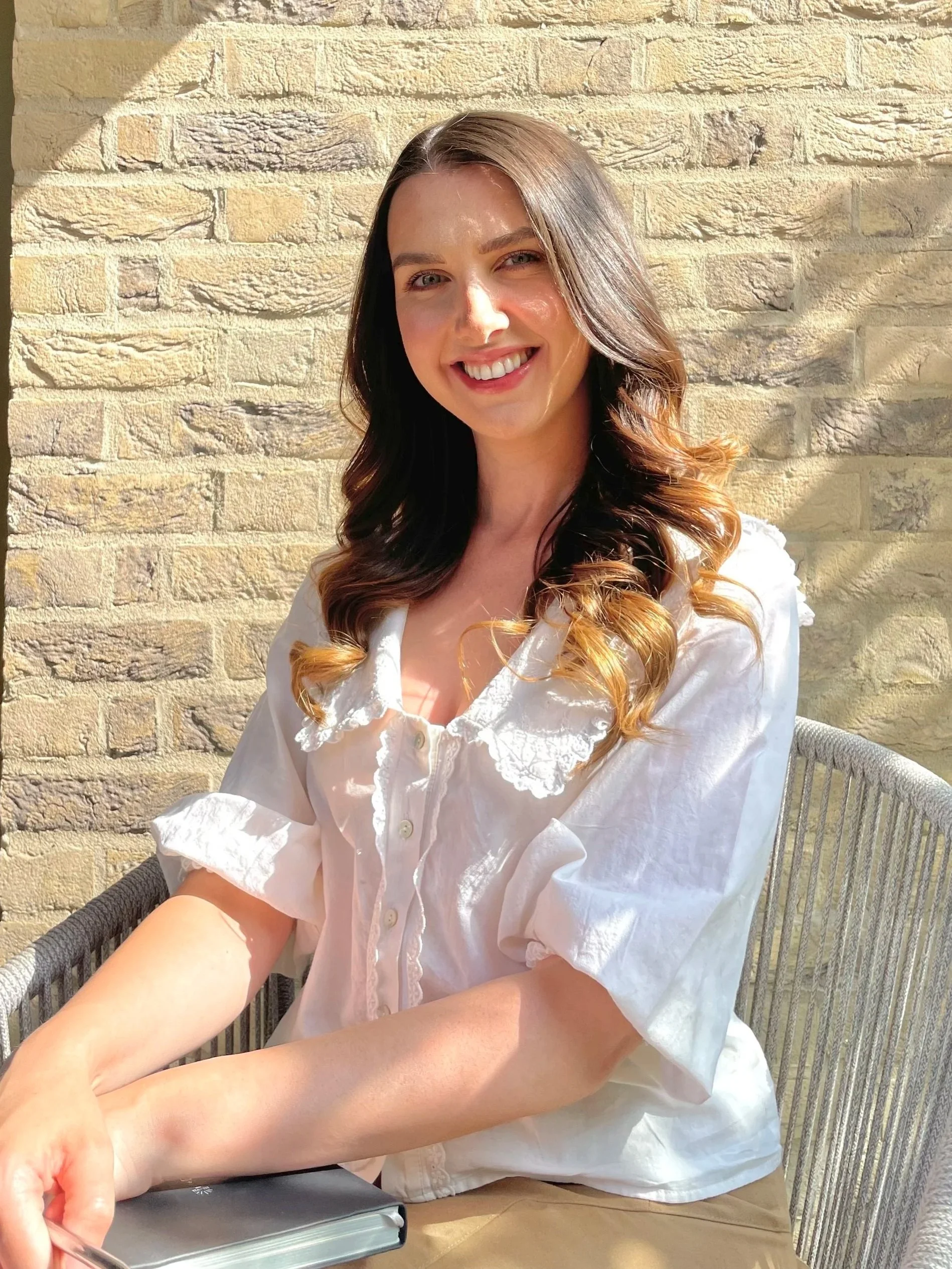 A young female therapist with long brown hair and a bright smile is sitting at a table outside in front of a brick wall, holding a pen and near a closed book or notebook.