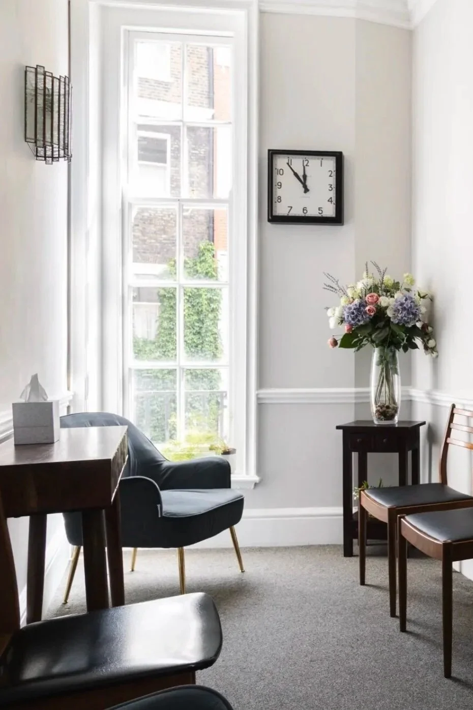 A room with a large window, a black armchair, a small side table with a vase of flowers, a wall clock showing 11:55, and two wooden chairs.