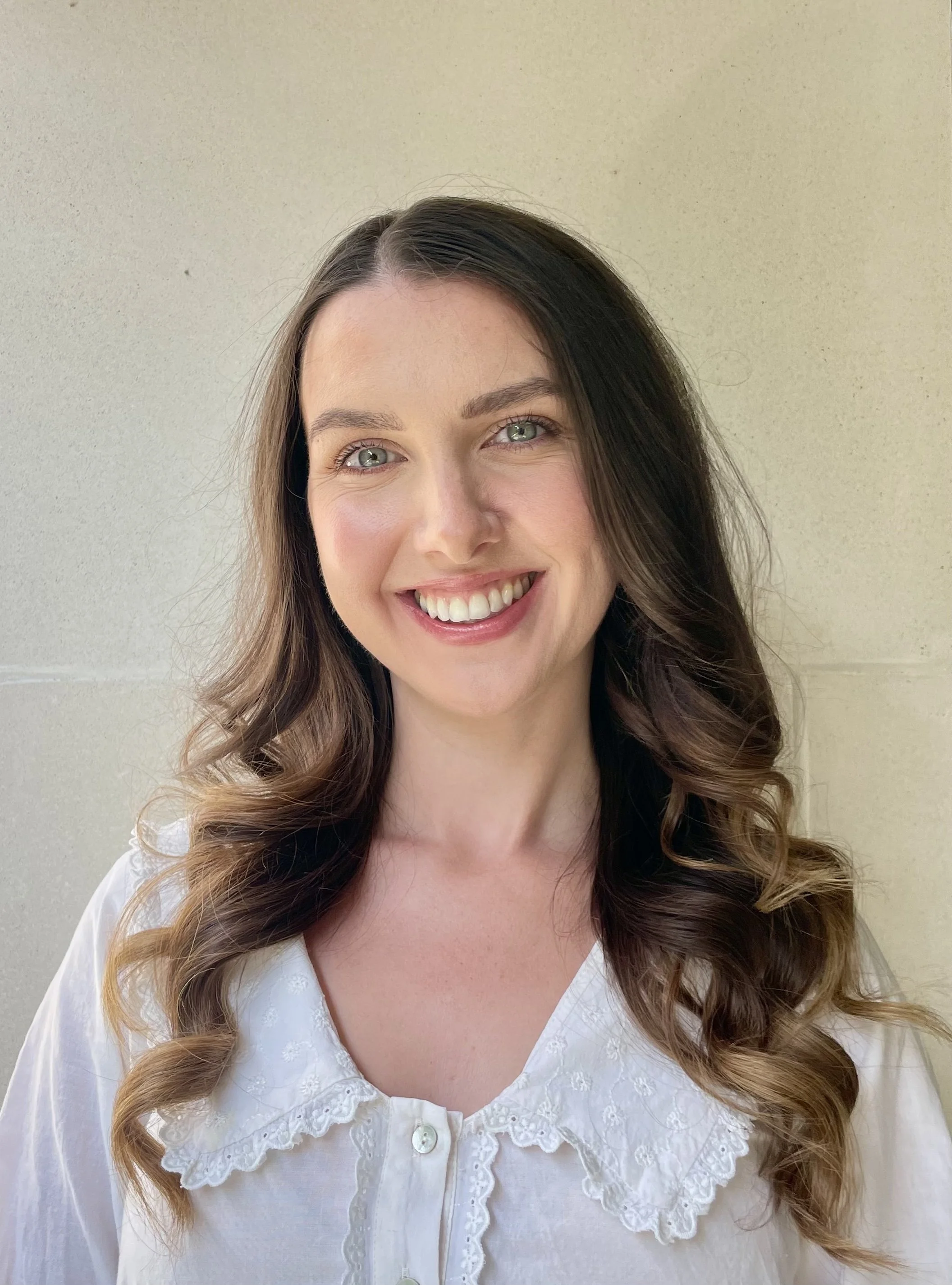 Close-up of a smiling therapist and practitioner health psychologist with long, wavy brown hair, wearing a white embroidered blouse, standing against a beige wall.