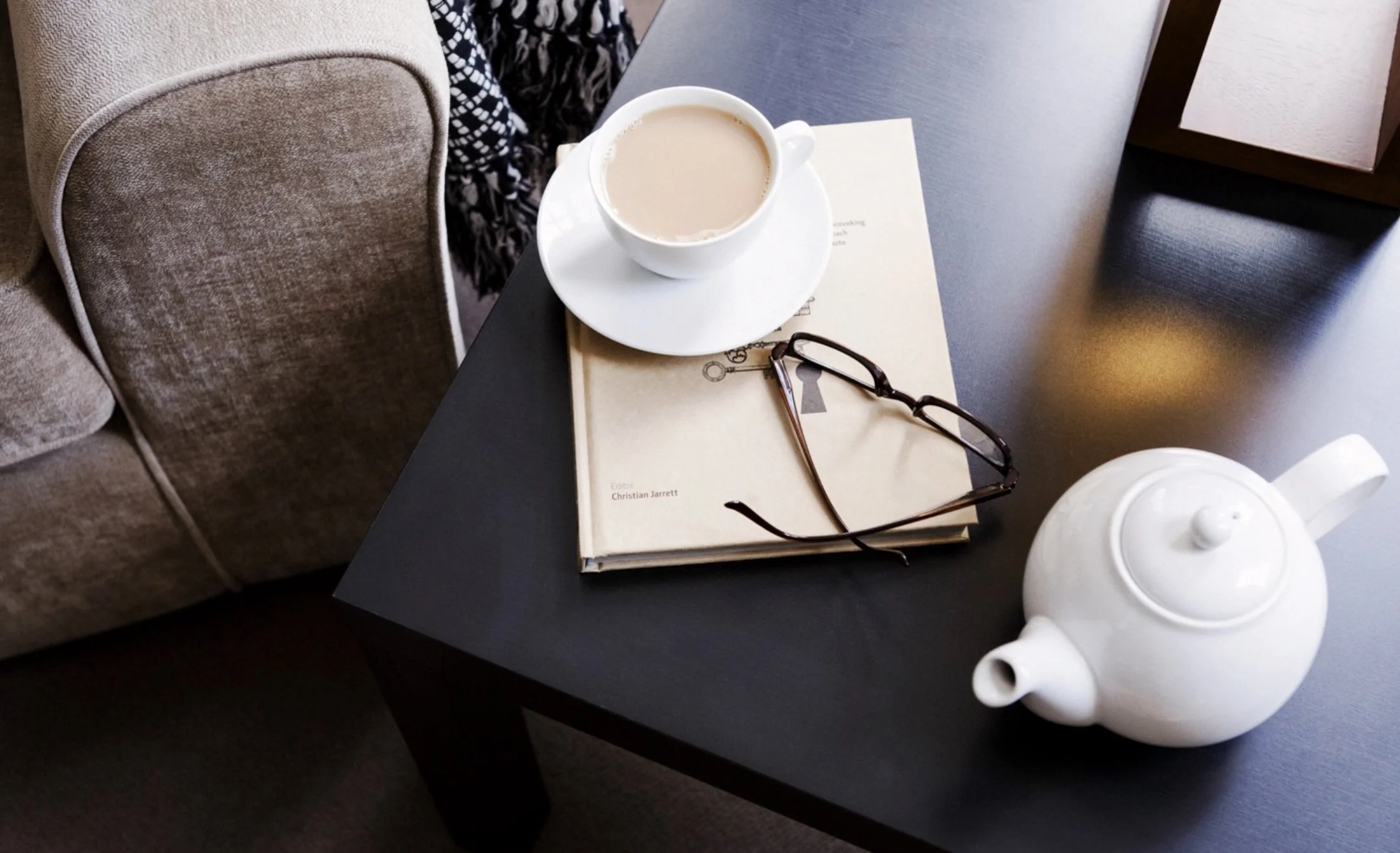 A black table with a white coffee cup on a saucer, a pair of black glasses, a beige book with the title 'Christian Jarrett', a white ceramic teapot, and a silver pen, next to a beige upholstered chair with a patterned scarf or cloth draped over its back.