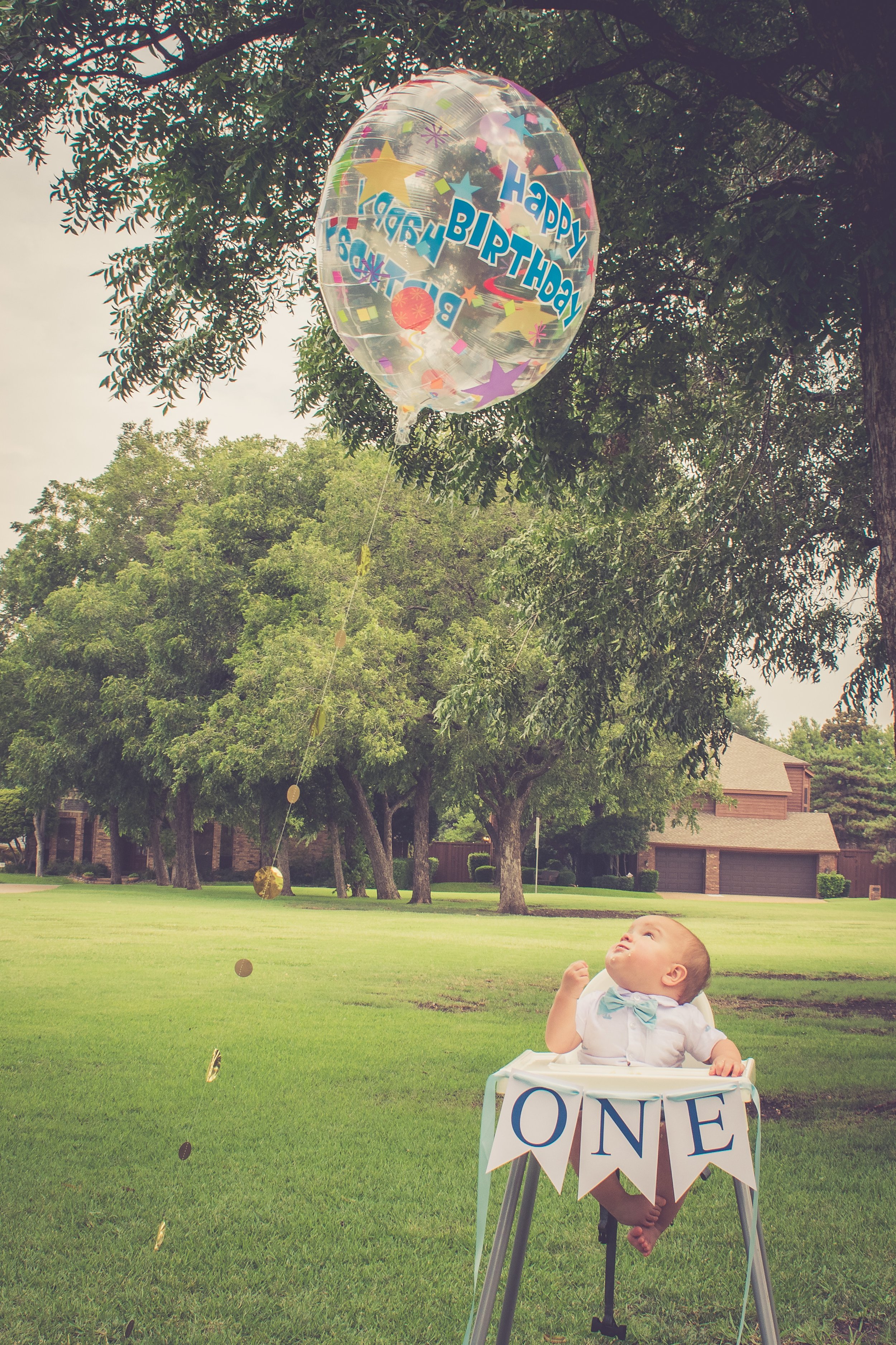 A young child in a high chair outdoors in a grassy yard, looking up at a clear balloon with colorful stars and the words 'Happy Birthday'. The child is holding a small object in one hand, and a sign with the word 'ONE' is hanging in front of the high chair.