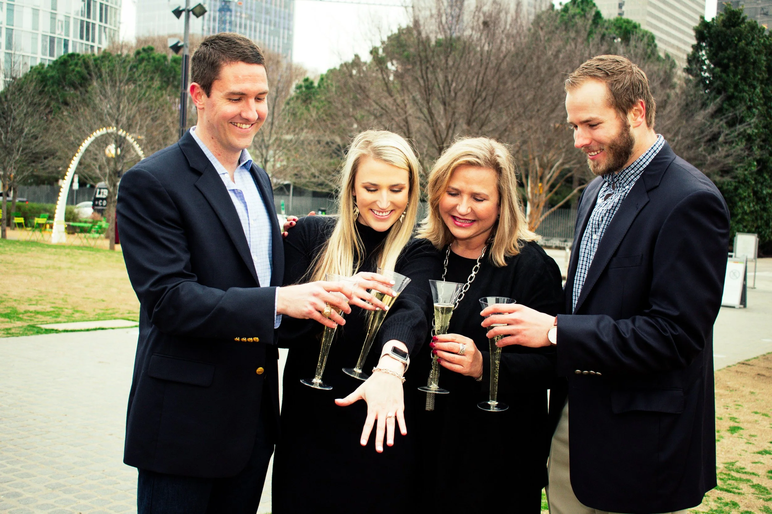 Four people at an outdoor event, dressed in formal attire, holding glasses of champagne and smiling.