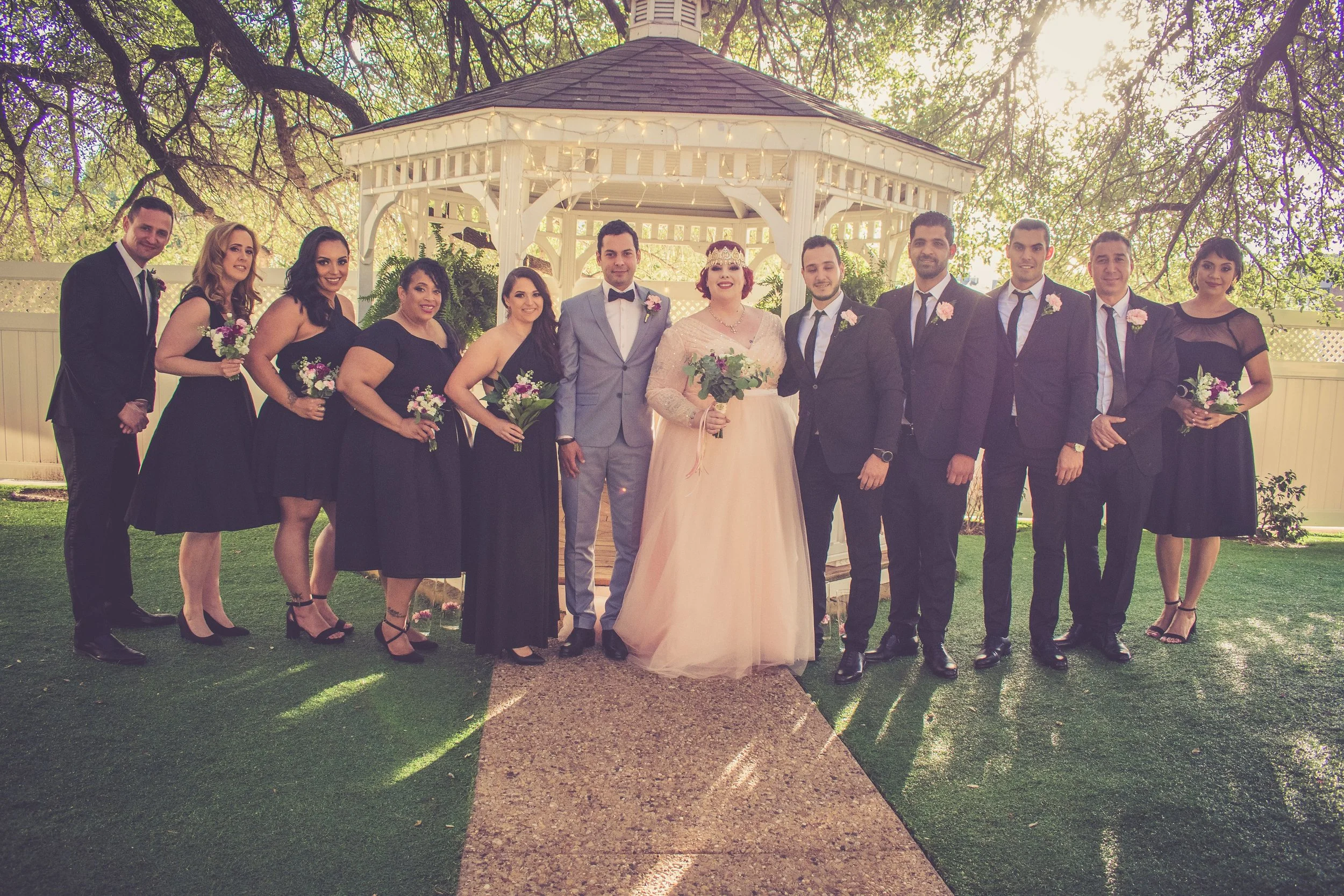 A group of people dressed in formal attire, standing in front of a gazebo with string lights during a wedding celebration outdoors.