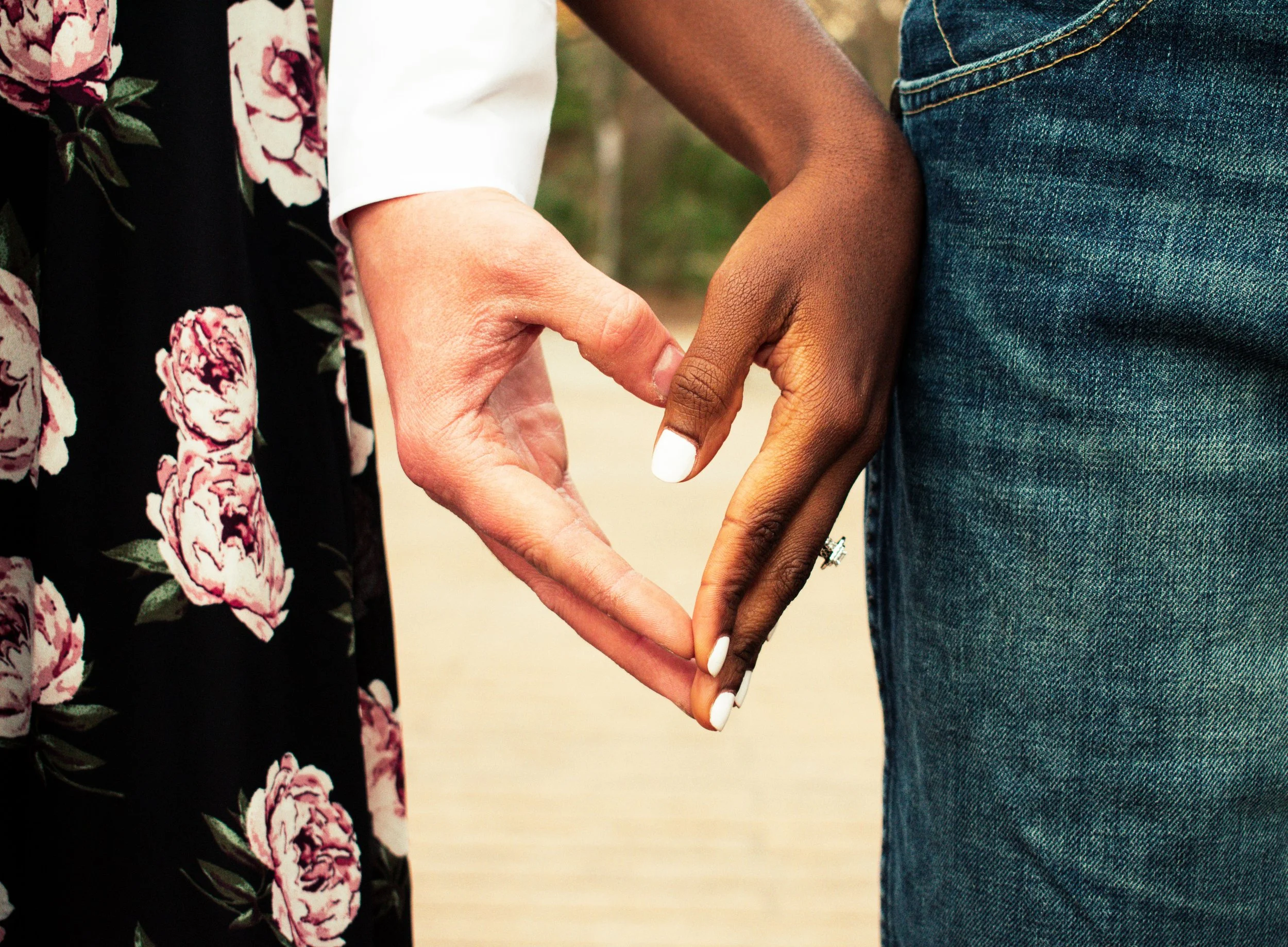 Two people holding hands in heart shape, one with lighter skin and white painted nails, wearing a floral patterned shawl, and the other with darker skin and denim jeans.