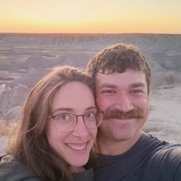 A smiling man and woman taking a selfie together outdoors during sunset with a scenic canyon in the background.