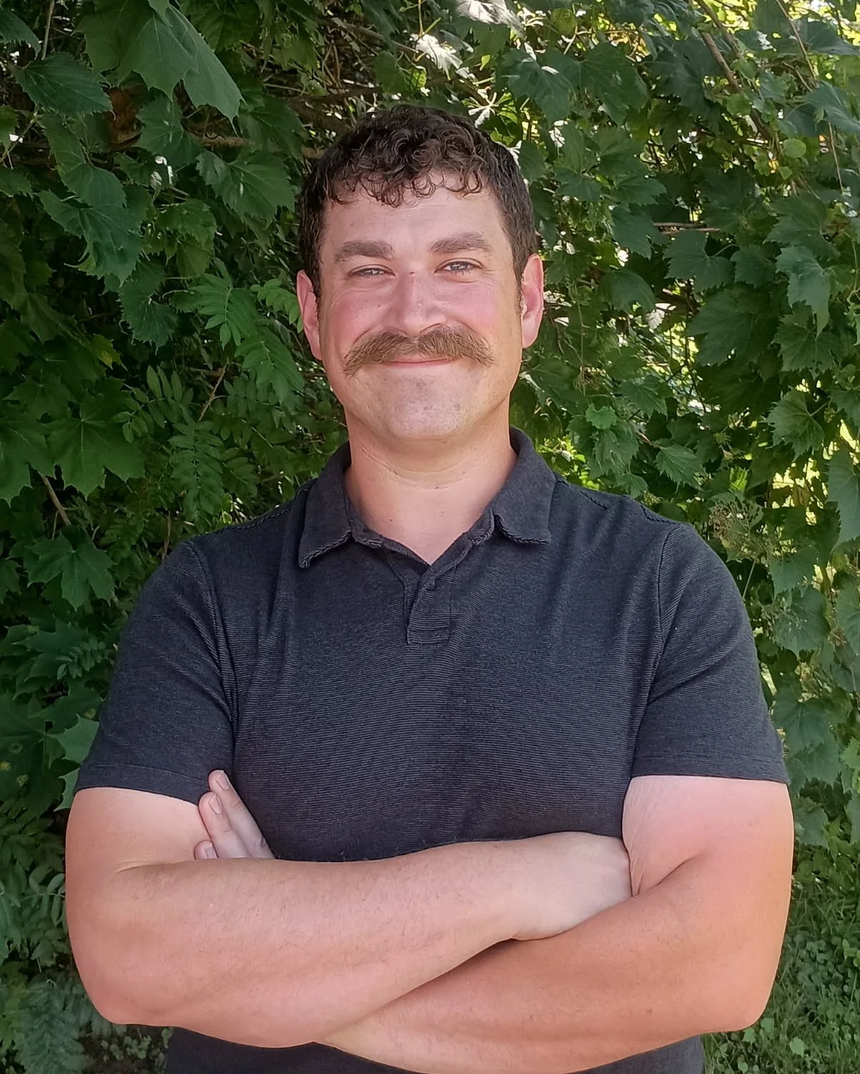A man with curly brown hair, a mustache, and a beard standing outdoors in front of leafy green trees, crossing his arms and smiling.