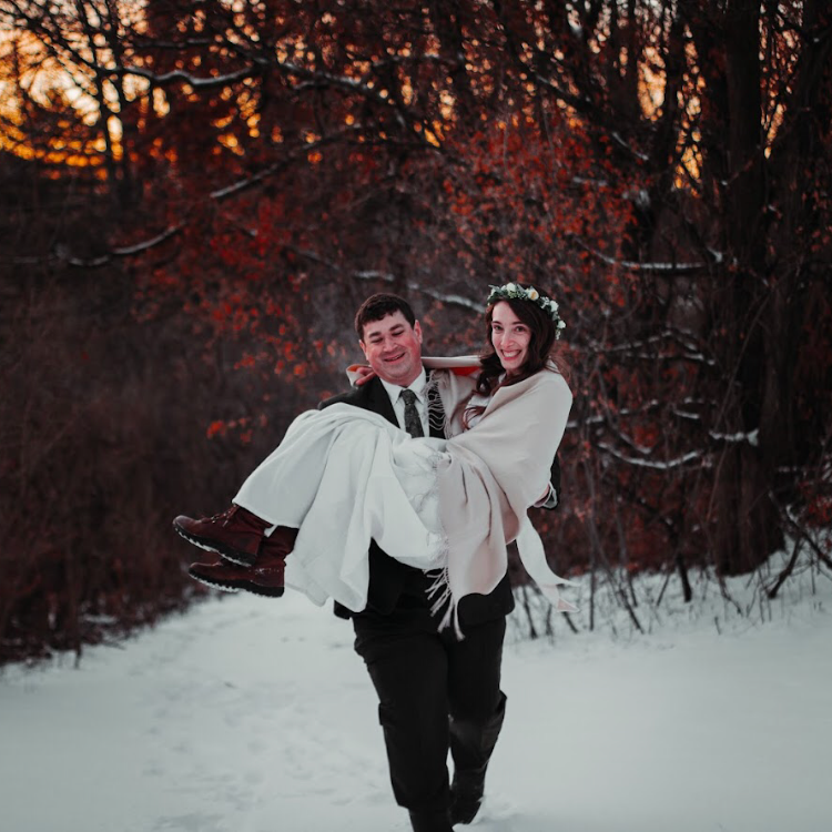 A smiling man in a suit carrying a smiling woman in a white dress and shawl, with a floral crown, in a winter outdoor setting with snow and red autumn trees in the background.