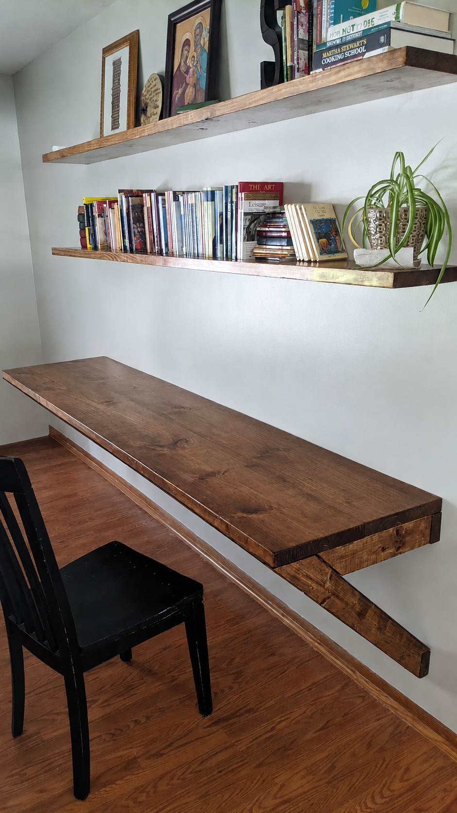 A wall-mounted wooden desk with two floating shelves above it, containing books, CDs, and decorative items including a plant in a white pot, religious icons, framed artwork, and a small decorative plate on a neutral wall with hardwood floors.