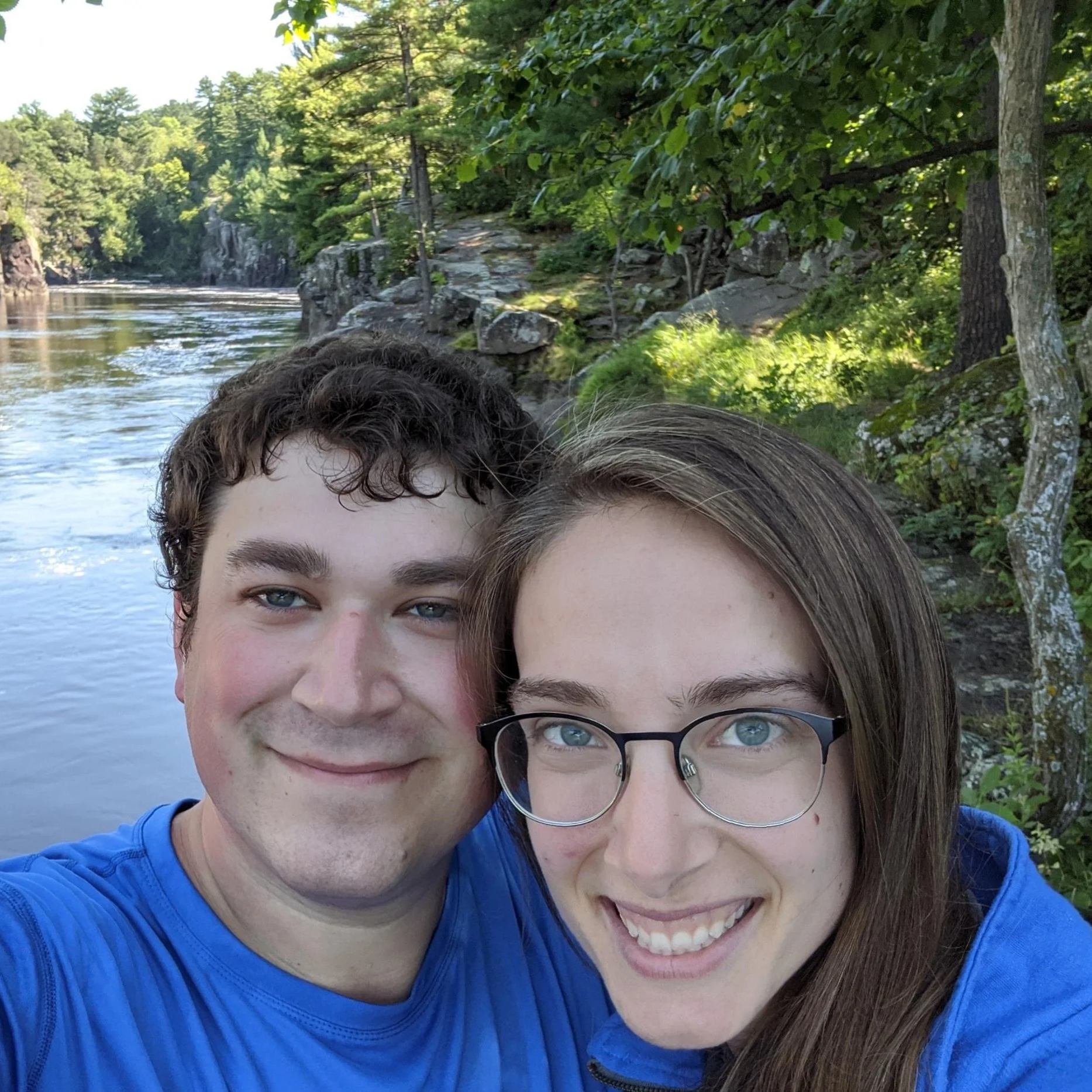 Close-up of two smiling people, a man with curly brown hair and a woman with straight brown hair and glasses, taking a selfie outdoors near a body of water with rocks and green trees.