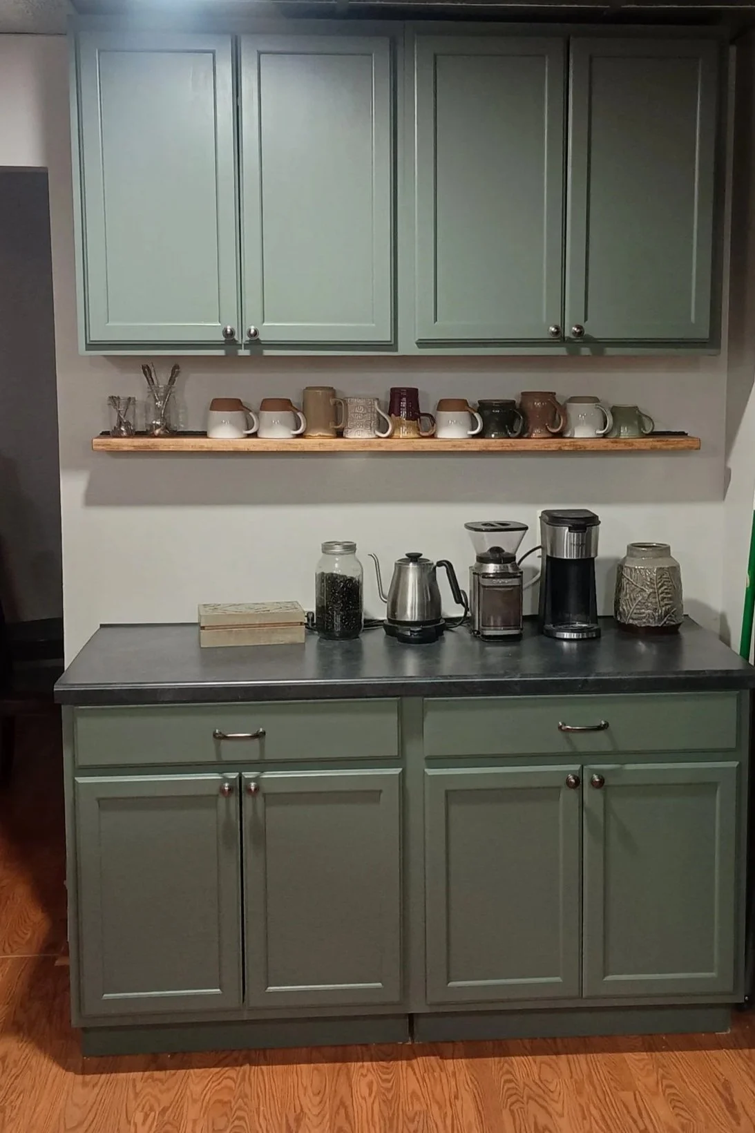A kitchen counter with a green cabinet, a wooden shelf holding mugs, and various coffee-making appliances, including a glass jar, an electric kettle, a coffee grinder, and a coffee maker.