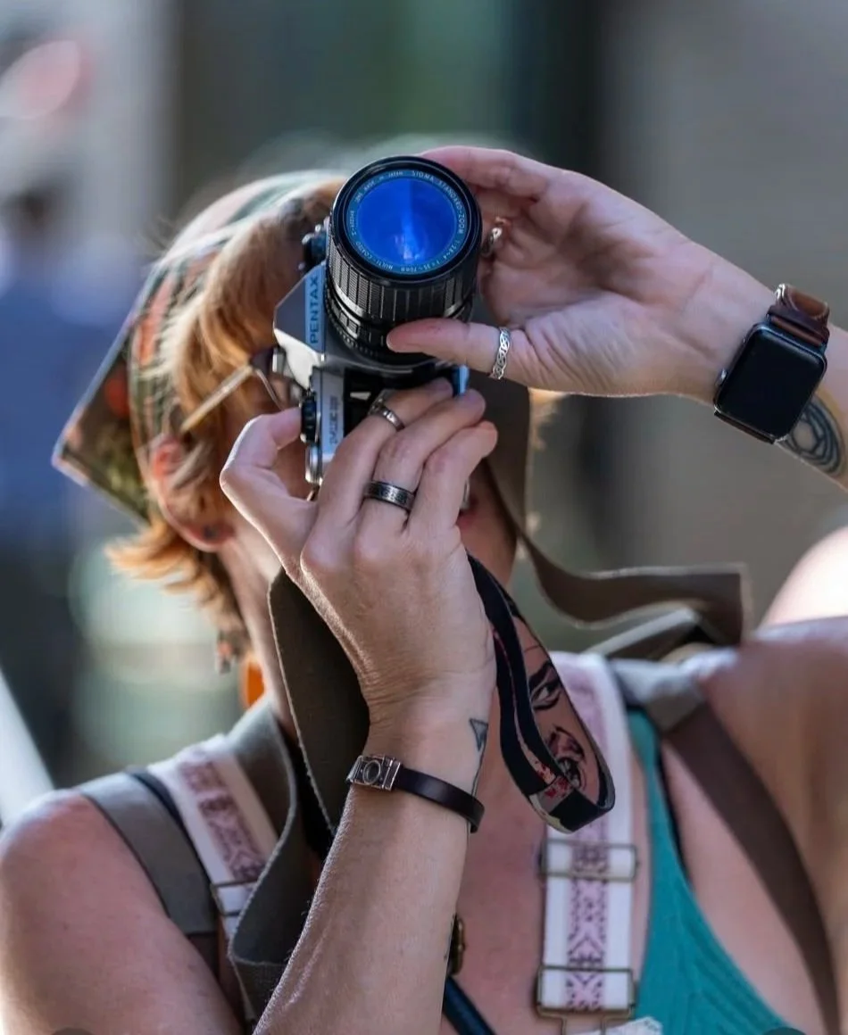 A person holding a vintage Pentax camera up to their face, wearing a sun hat, rings, bracelets, and a smartwatch, with an outdoor background.