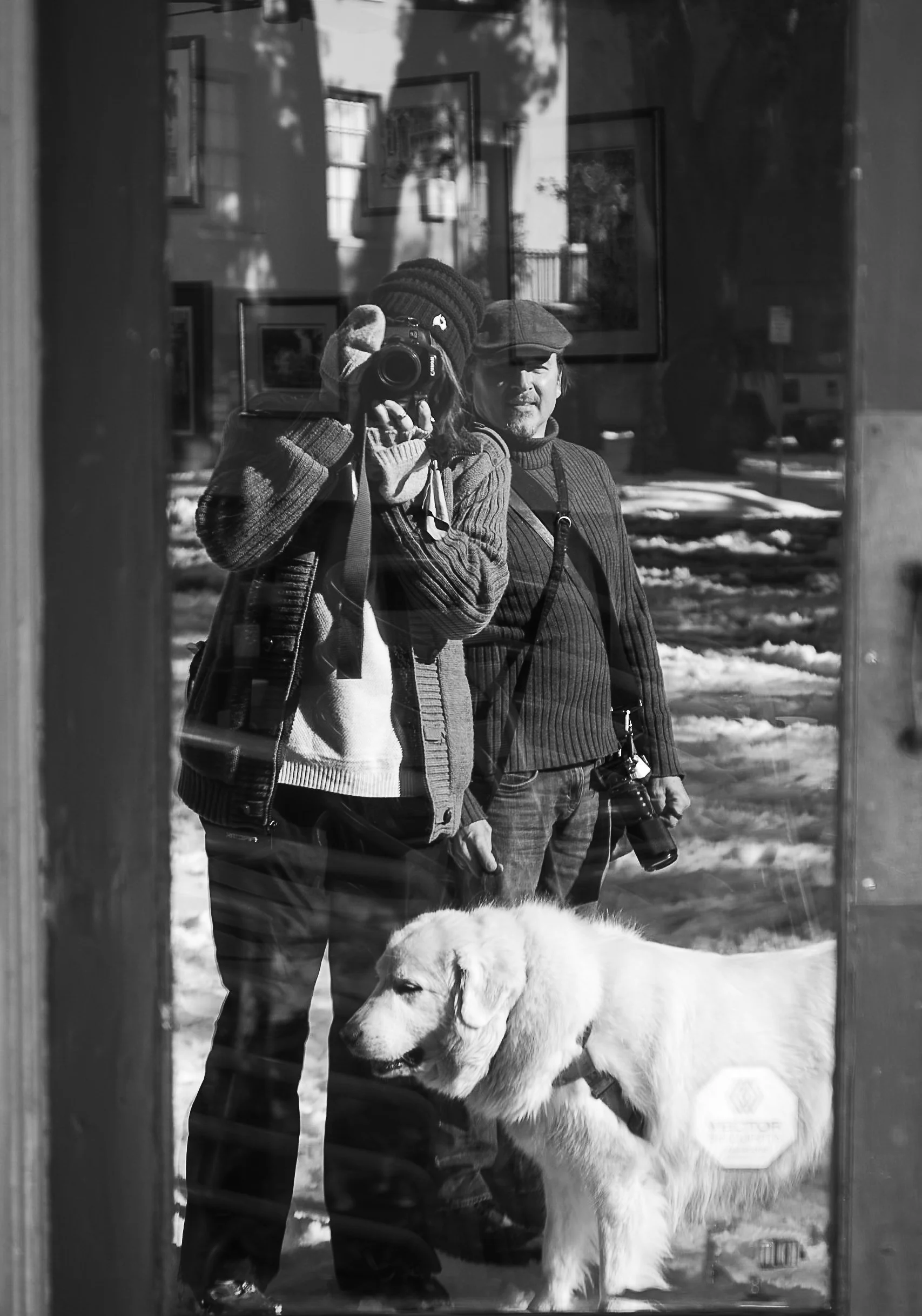 Reflection of a person taking a photo with a dog next to them, standing on snow in front of a building. The person is wearing a beanie, gloves, and sweater; the dog is a Great Pyrenees