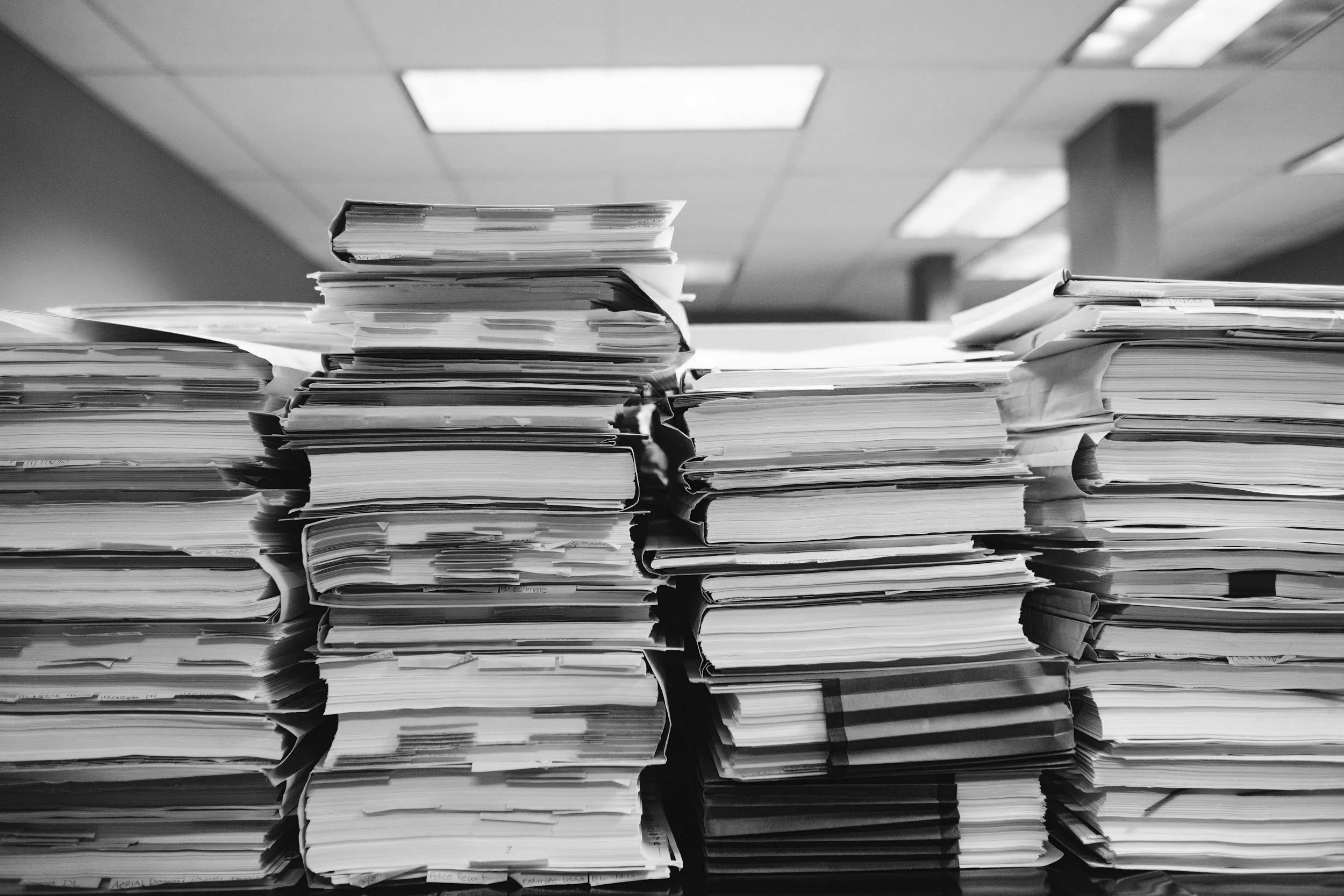 Schwarze und weiße Aktenordner und Papierstapel auf einer Bürobürotischplatte, im Hintergrund eine Bürodecke mit Deckenleuchten.