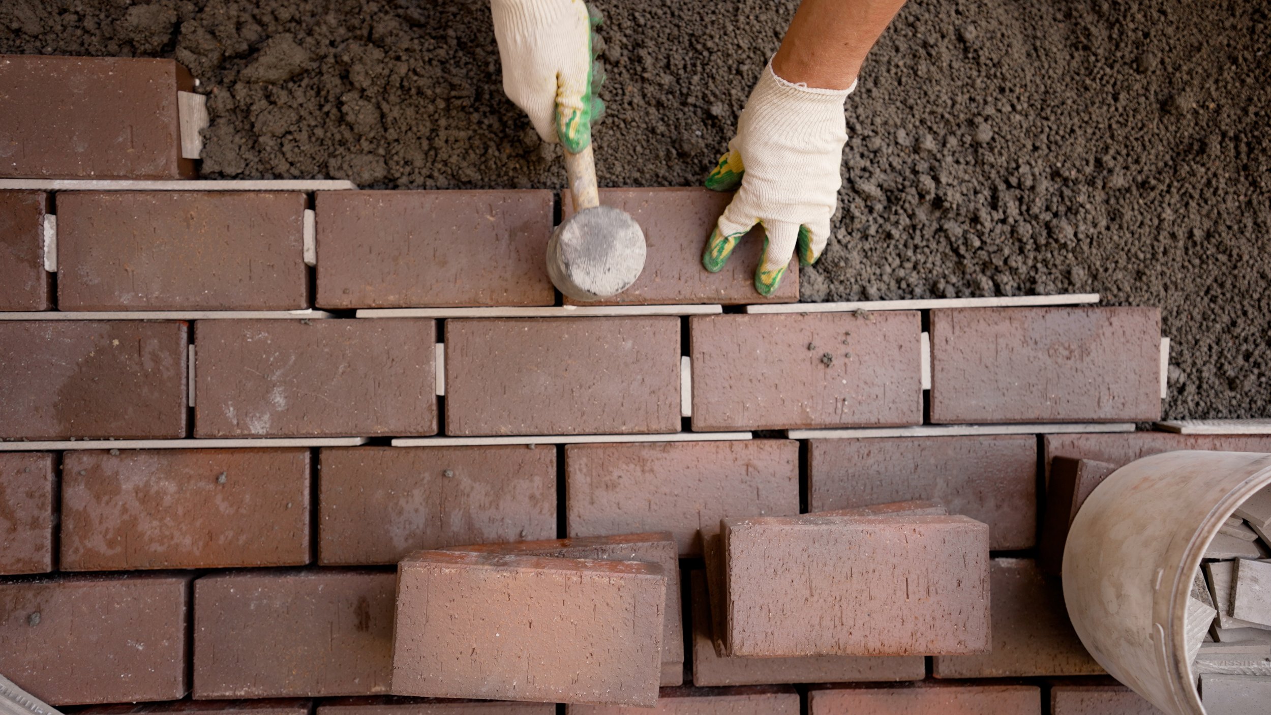 Person laying bricks, using a hammer and wearing gloves.