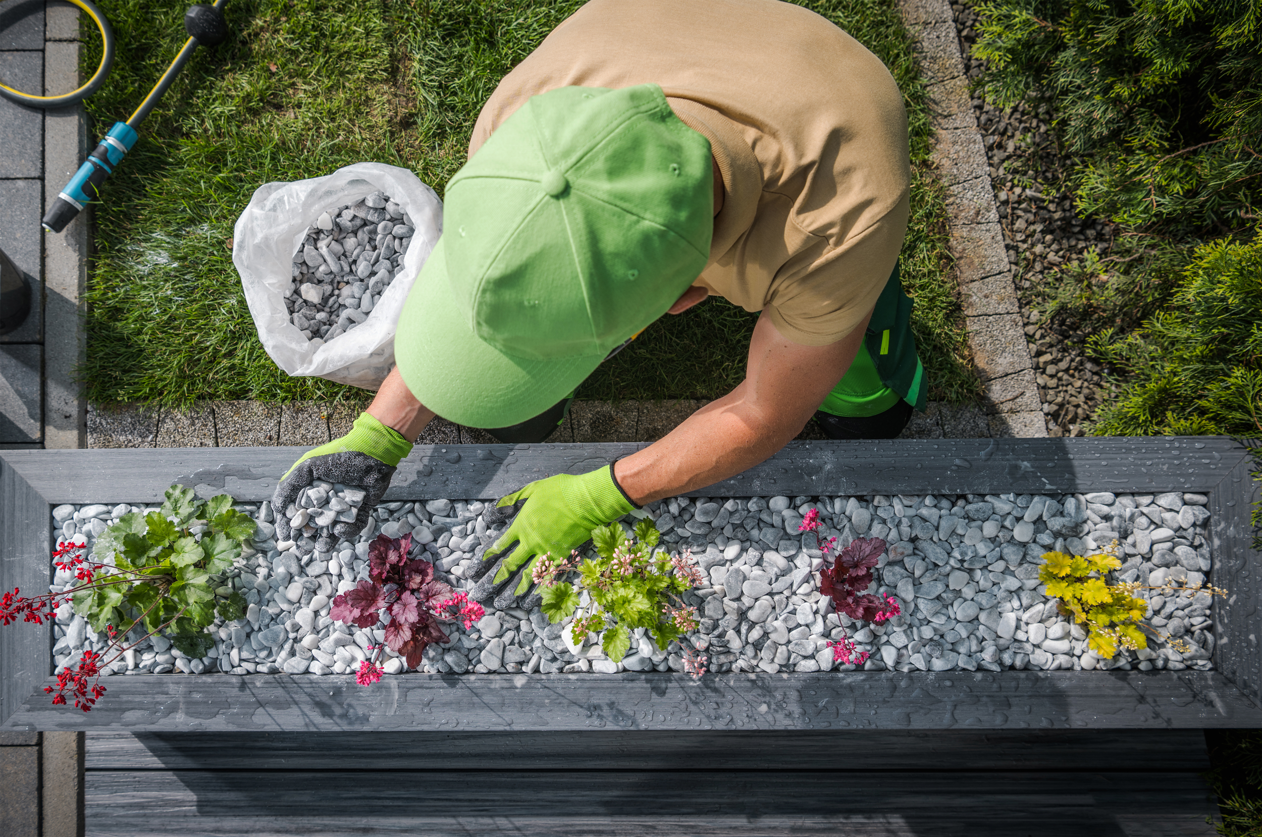 Person planting flowers in a raised garden bed filled with gray rocks, wearing a green hat and gloves.