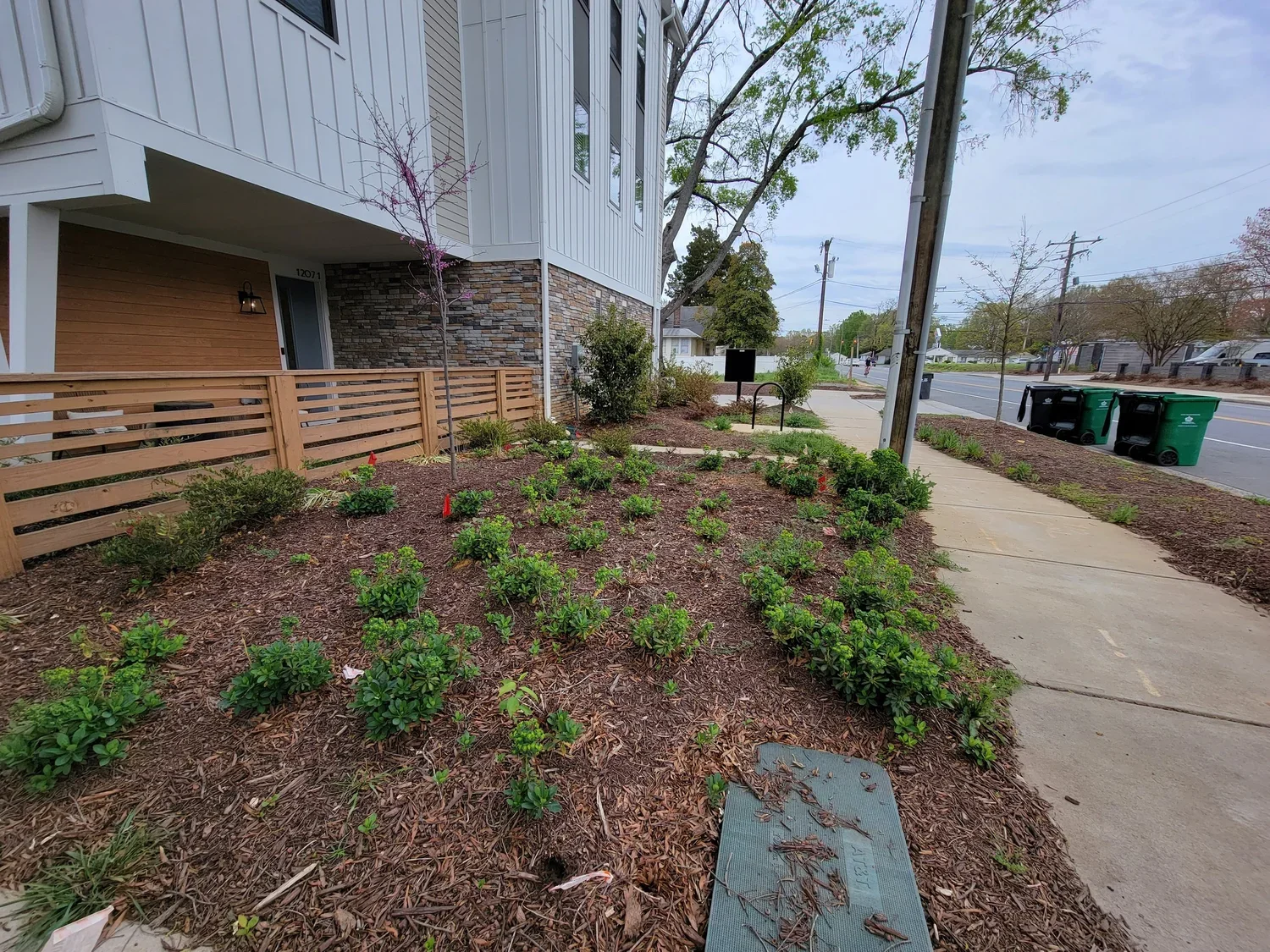 Residential front yard shrubs and small trees, a wooden fence, sidewalk, street with trash bins, utility poles, and a multi-story house with stone and siding exterior.